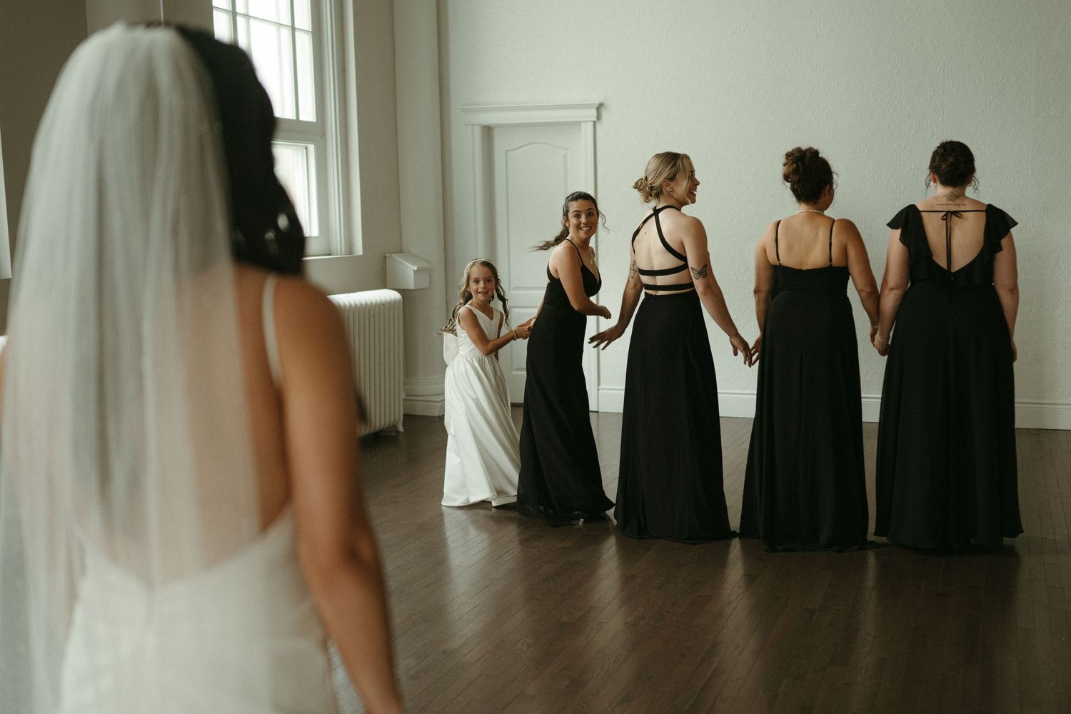 Bride faces bridesmaids in black gowns and flower girl during an emotional first look reveal in white room.