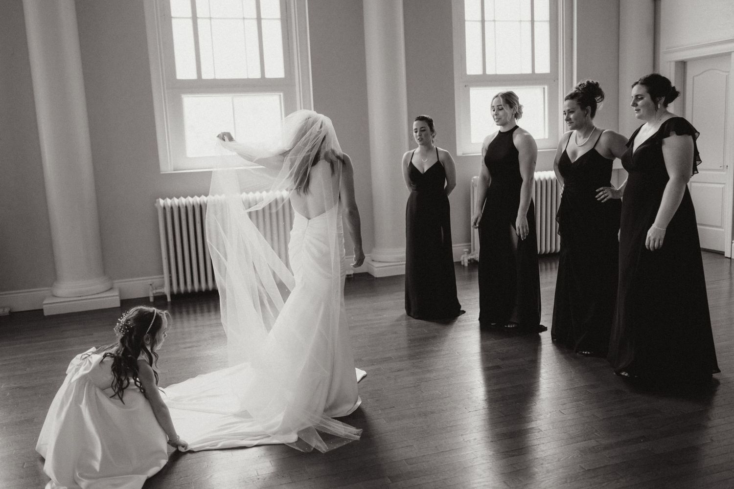 Bride with bridesmaids in black dresses during wedding preparations in black and white photo.