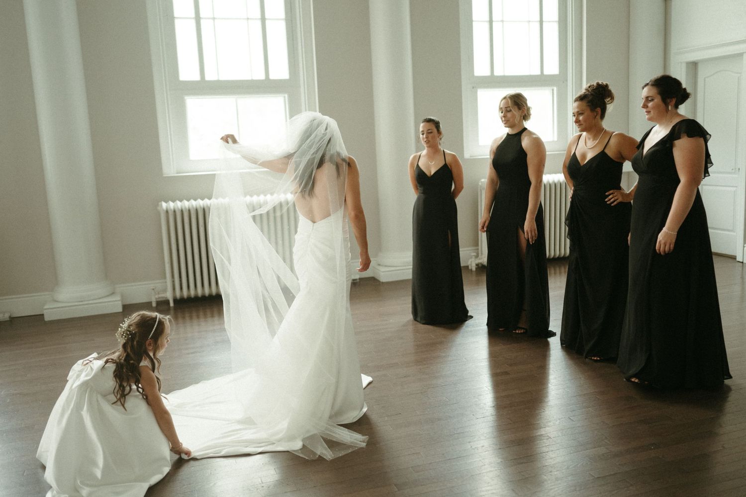 Bride in white gown with veil surrounded by bridesmaids in black dresses in a bright indoor venue.