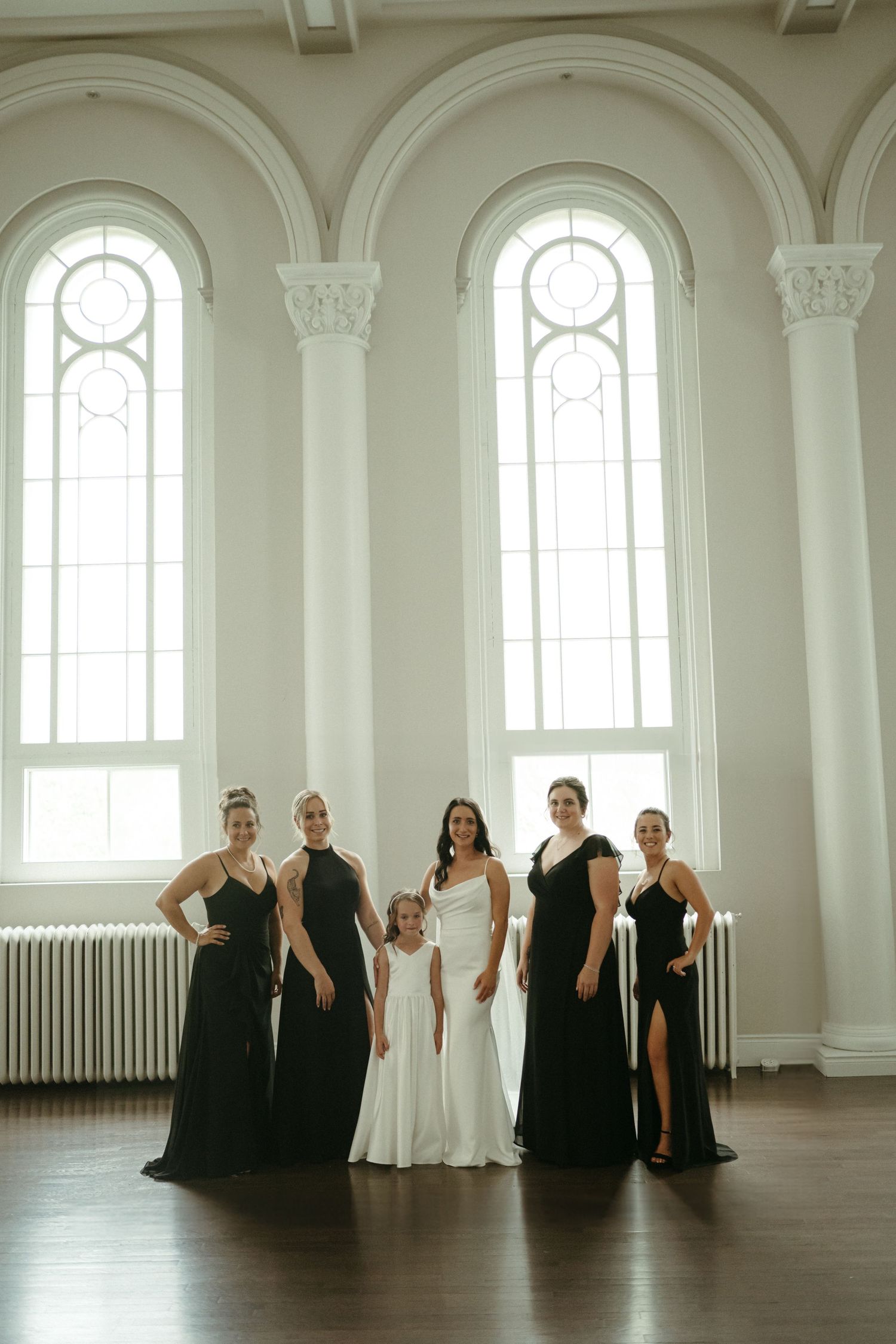 Bride in white gown poses with bridesmaids in black dresses and flower girl in bright hall.