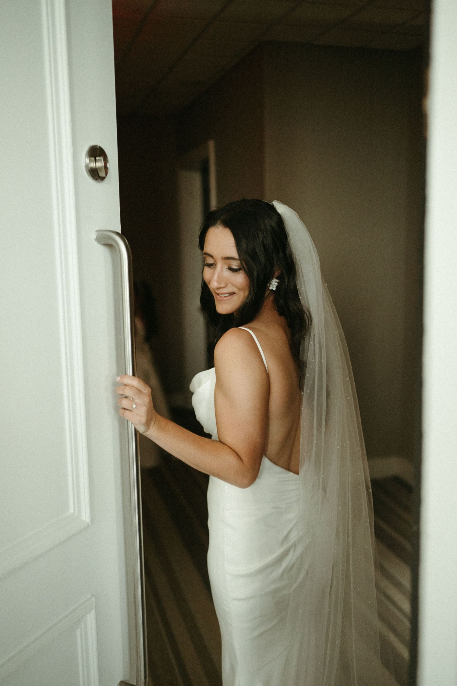 Bride in elegant backless white gown and veil smiles softly while holding a door at wedding venue.
