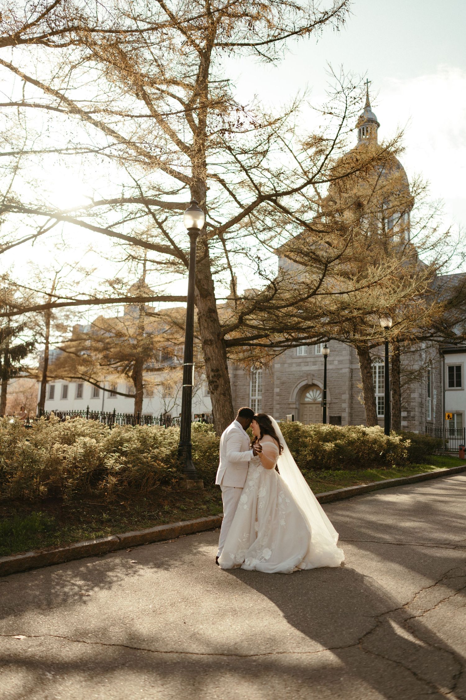 A bride and groom share a romantic embrace on a sunlit path beside a historic church surrounded by autumn trees.