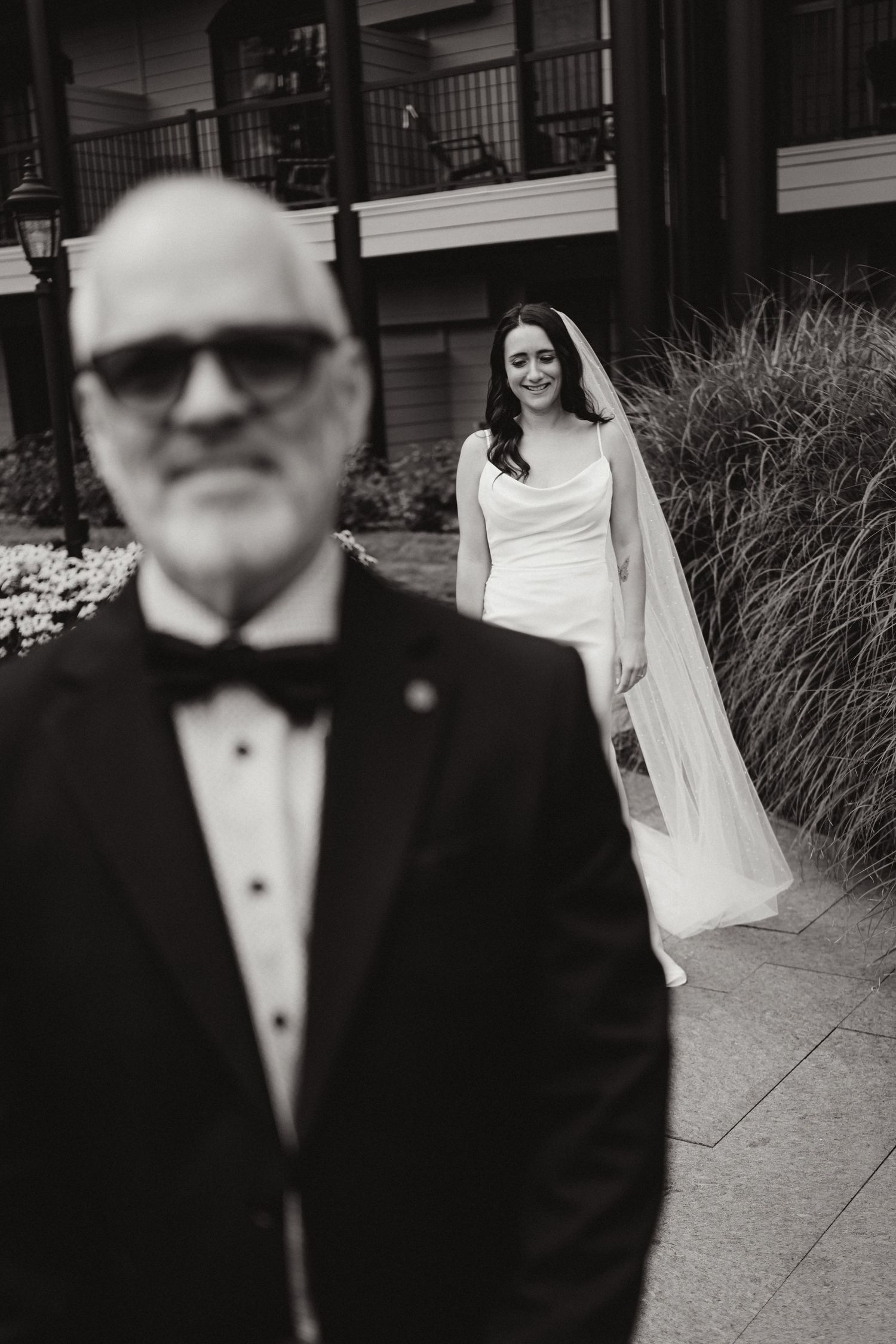 Black and white photo of a bride in a white gown and veil smiling as an older man in a tuxedo stands in the foreground.