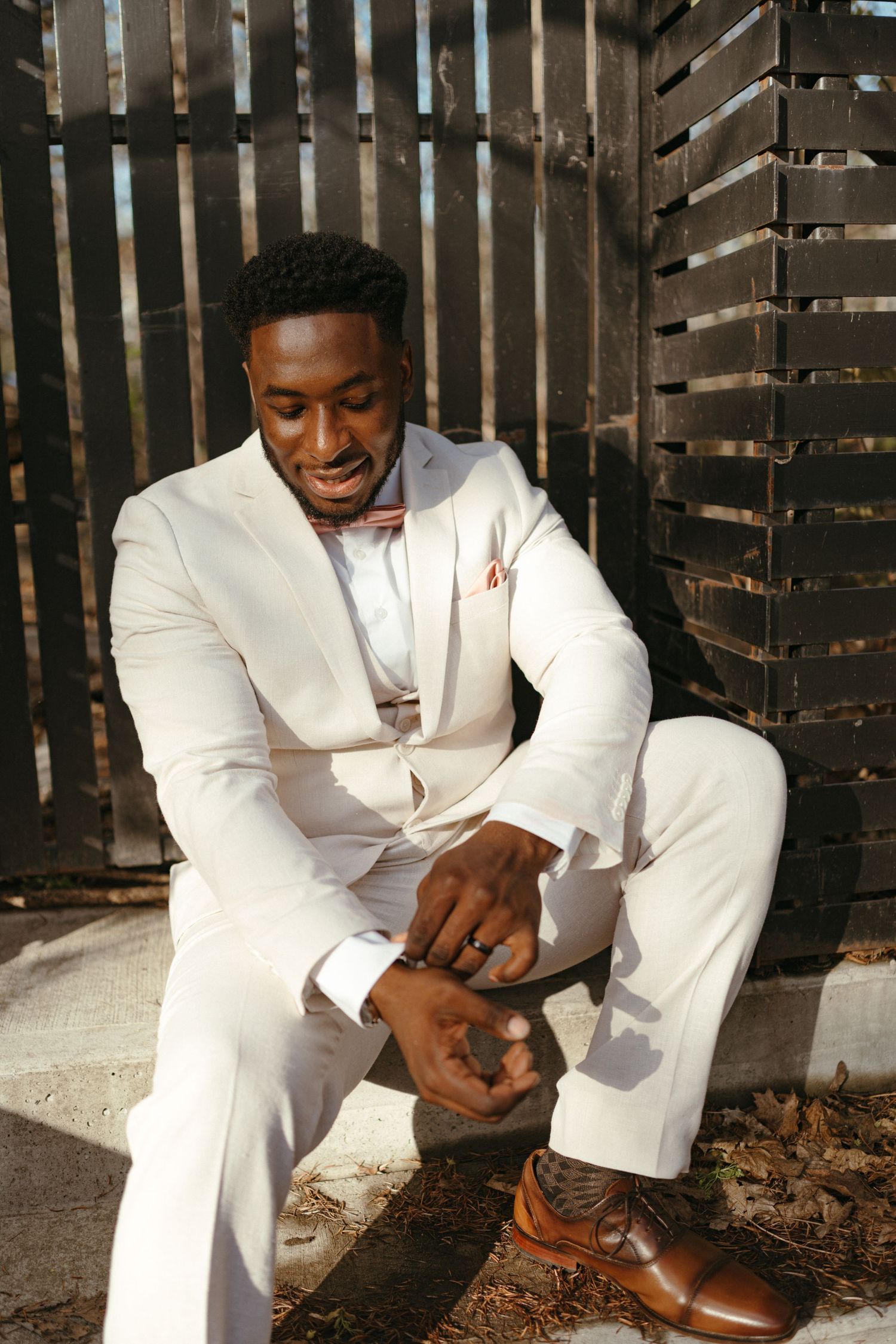 Dapper groom in white suit and bow tie sitting outdoors adjusting his brown dress shoes.