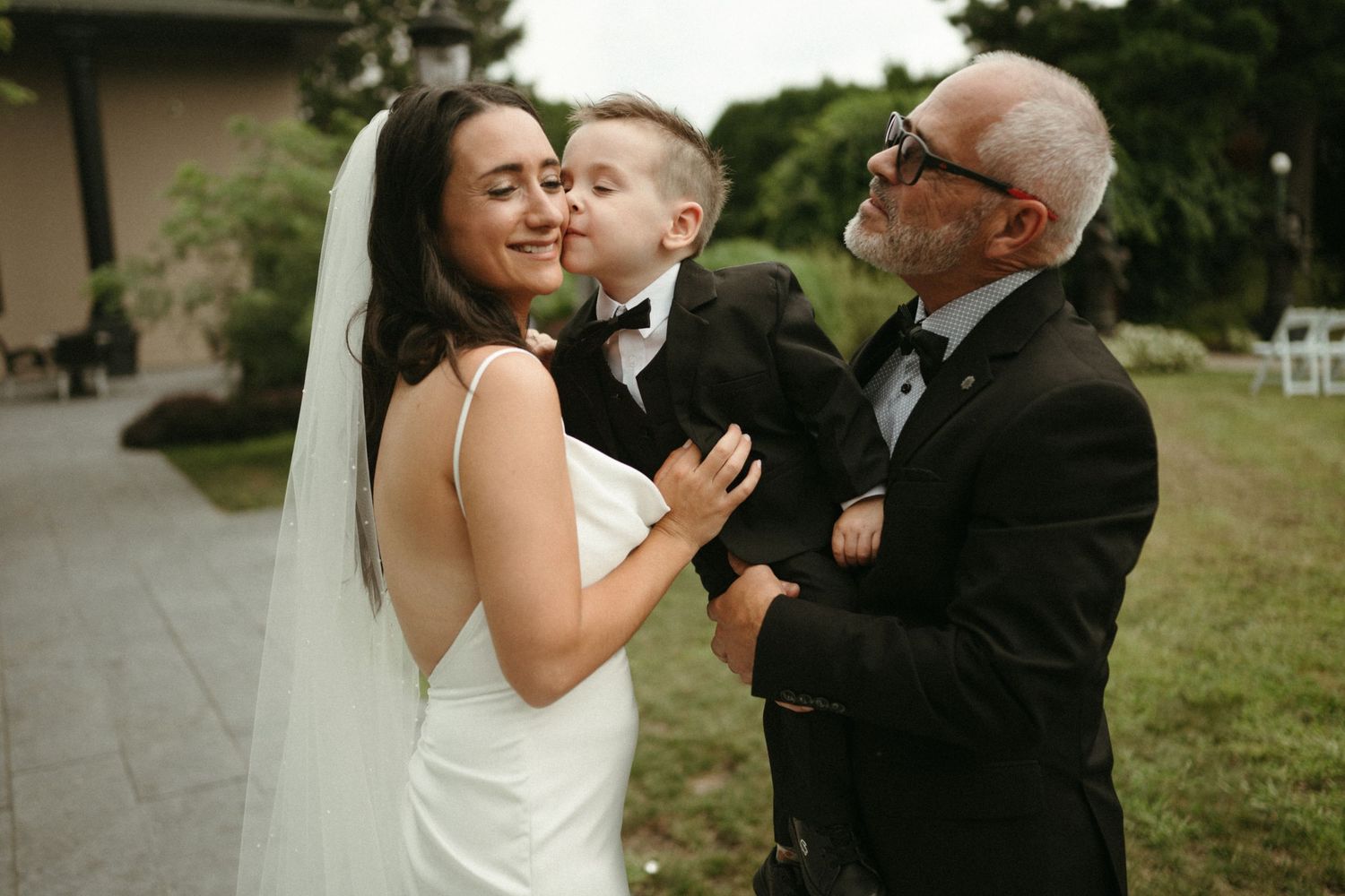 Bride in white gown receives sweet kiss from young boy in tuxedo while older gentleman looks on outdoors.