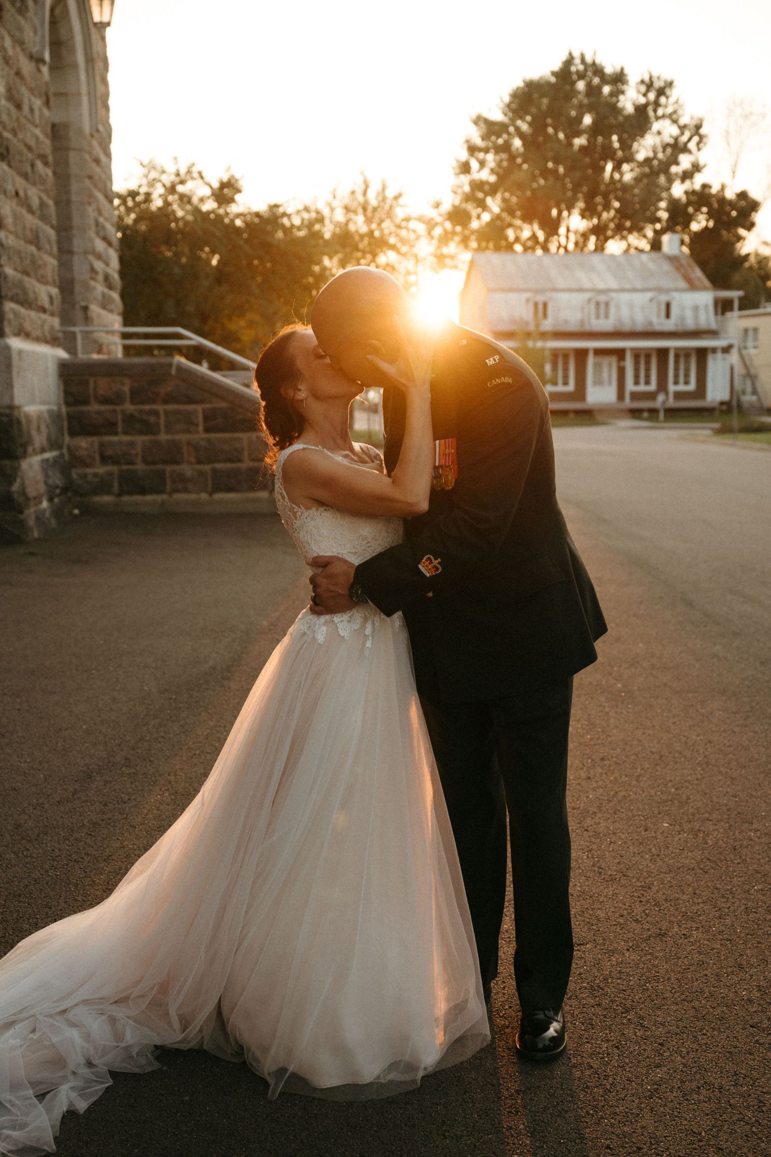Bride and groom share romantic sunset kiss, golden backlight glowing between them on wedding day.