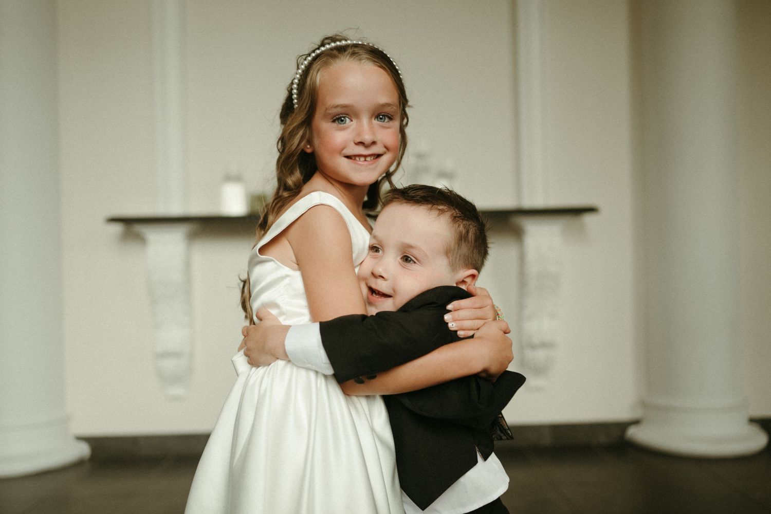 Flower girl in white dress hugs young ring bearer in black tuxedo at wedding reception.