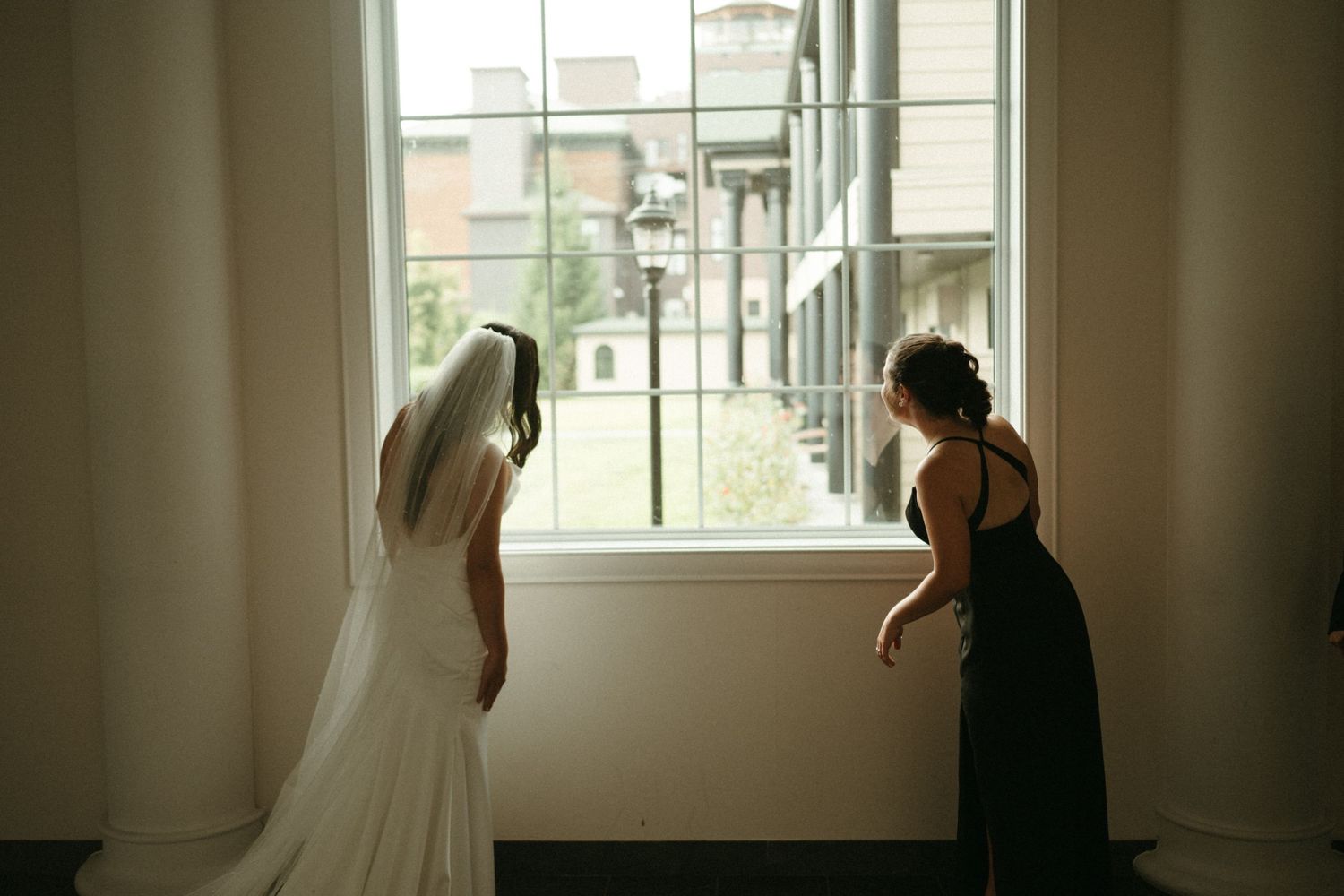 Bride in white gown and veil standing at window with bridesmaid in dark dress beside her.