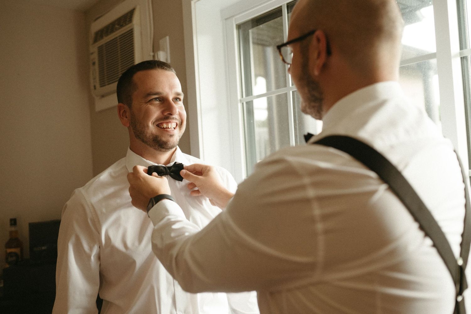 A man smiling as his groomsman helps him put on a black bow tie before a wedding ceremony.