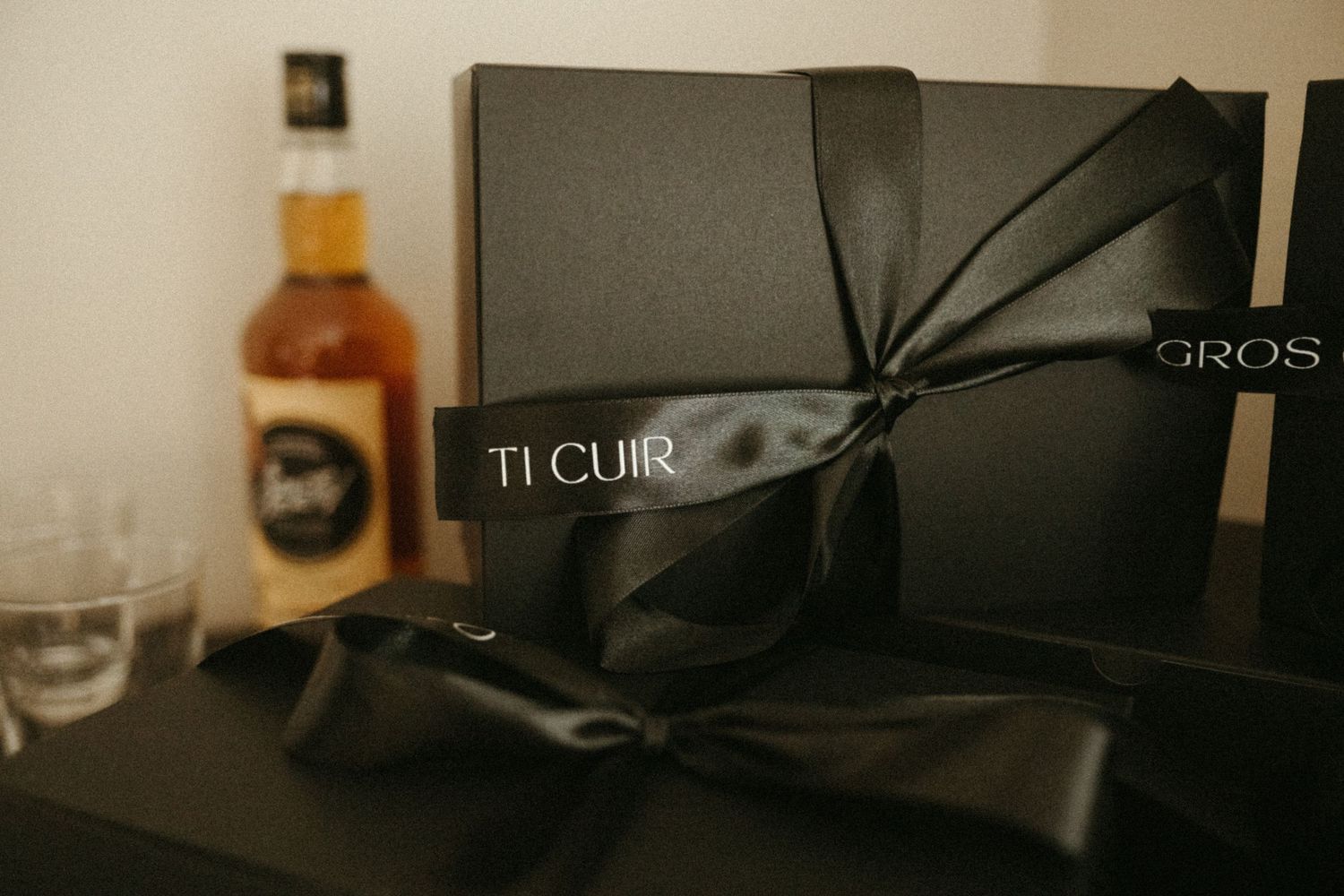 Elegant black gift boxes tied with satin ribbons labeled Ti Cuir and Gros beside a whiskey bottle.