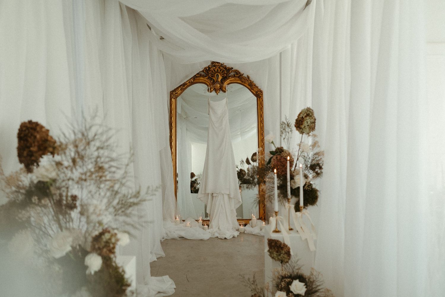 Elegant bridal gown displayed near ornate gold mirror with white draping and dried floral arrangements.