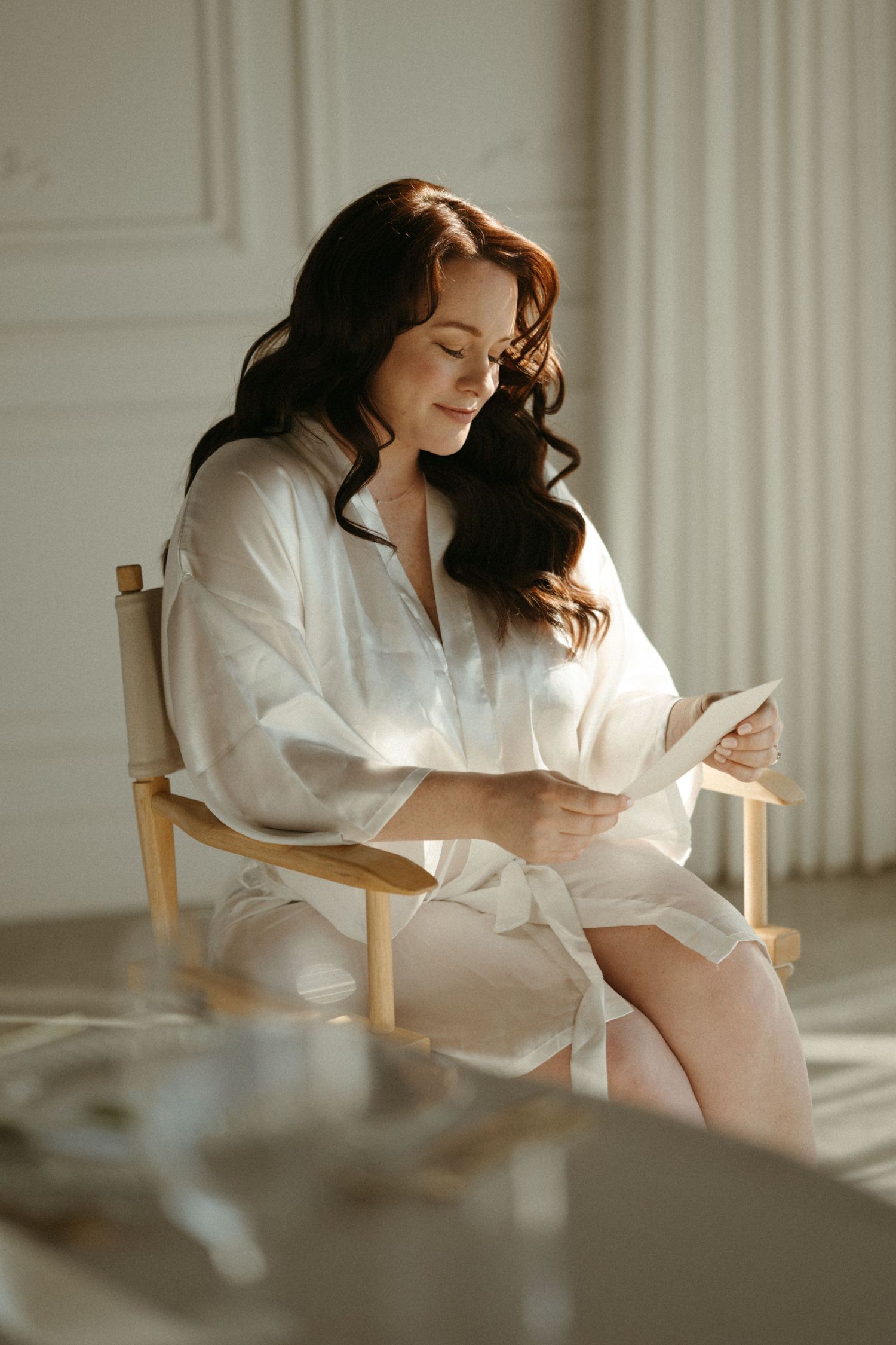 Bride in white robe sits in wooden chair reading a card, smiling softly in warm natural light.