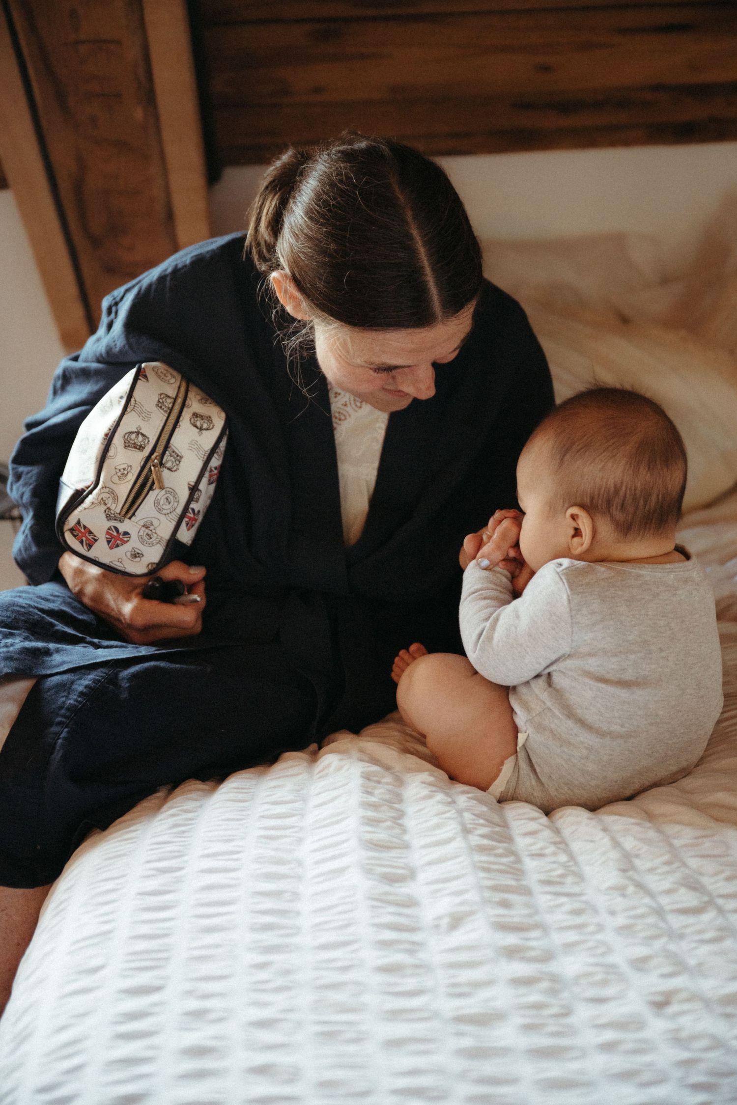 A mother in a black blazer sits on a white bed smiling at her baby in a gray onesie.