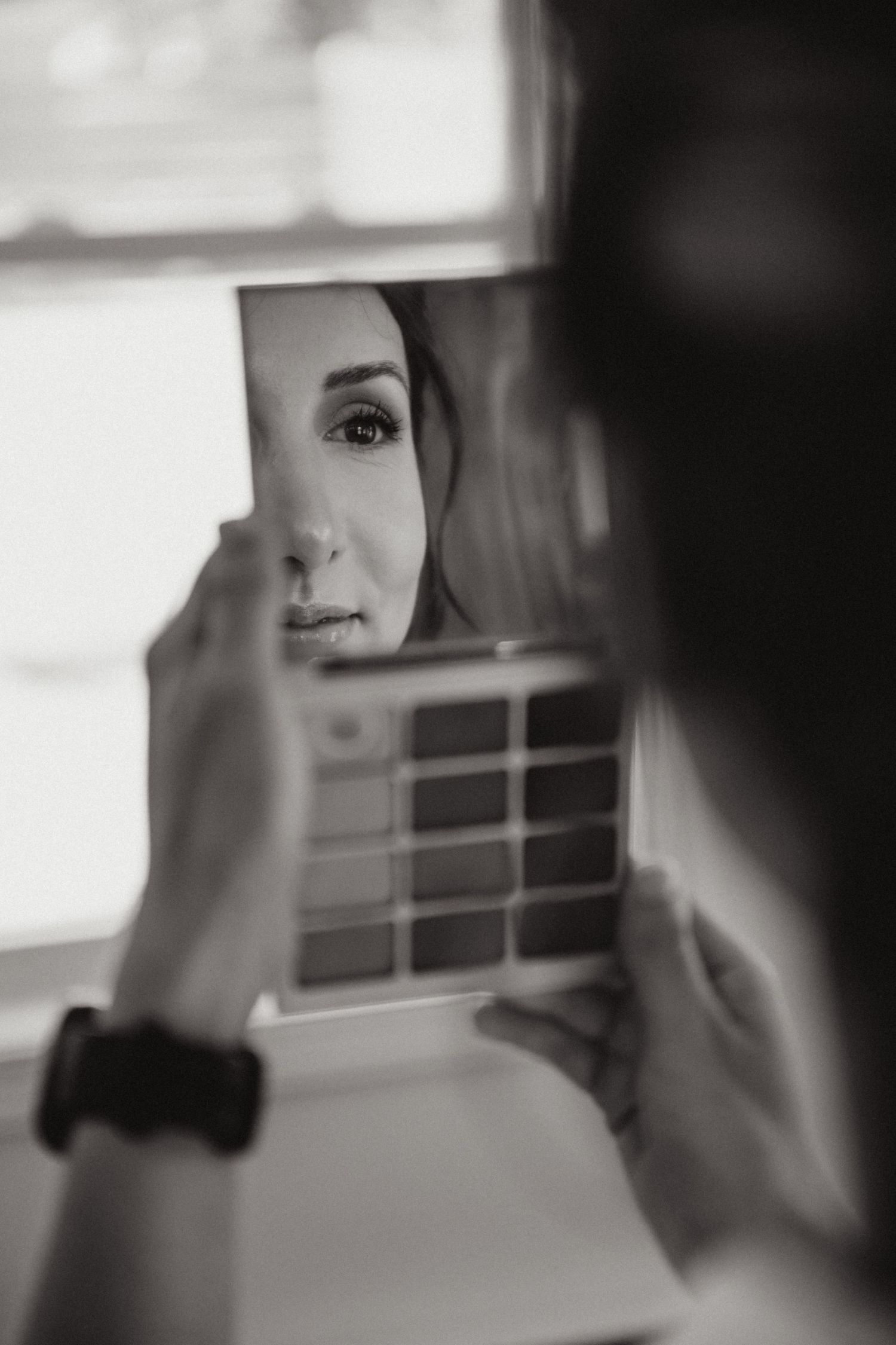 Black and white photo of a bride examining her makeup in a small mirror before her wedding.