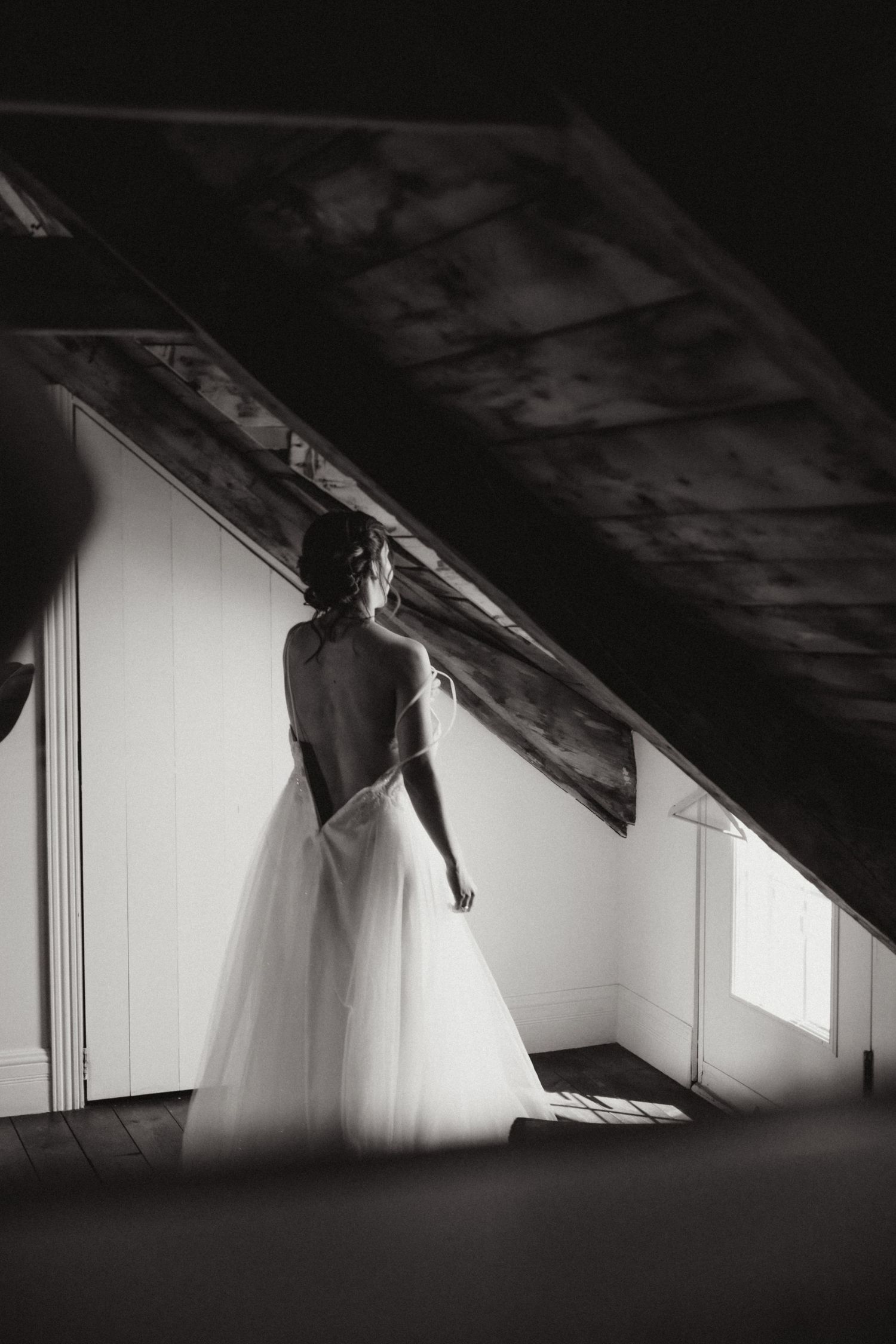 Bride in backless white tulle gown stands in dramatic black and white attic light.