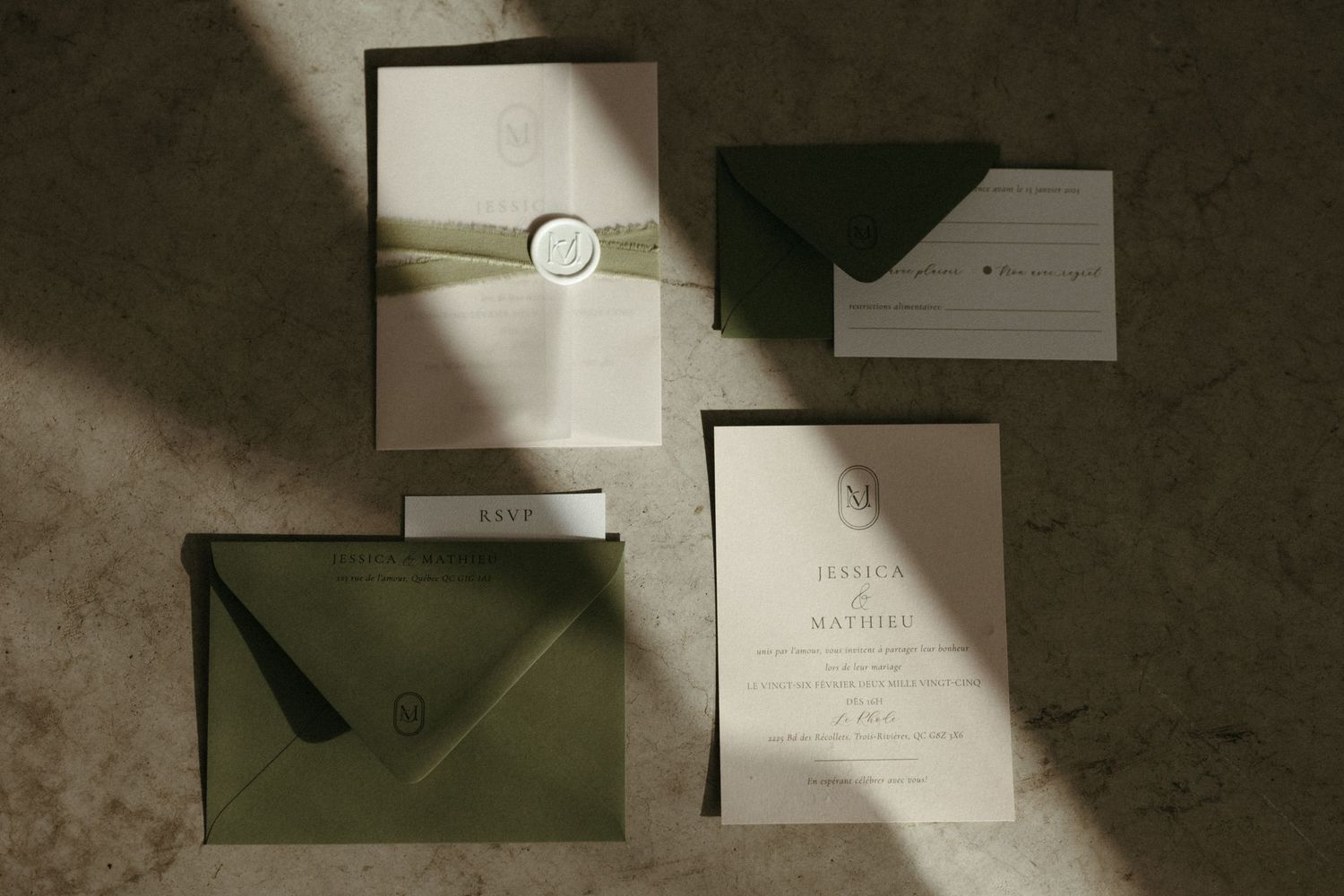 Elegant wedding invitation suite with sage green envelopes, wax seal, and ribbon on a neutral stone surface.