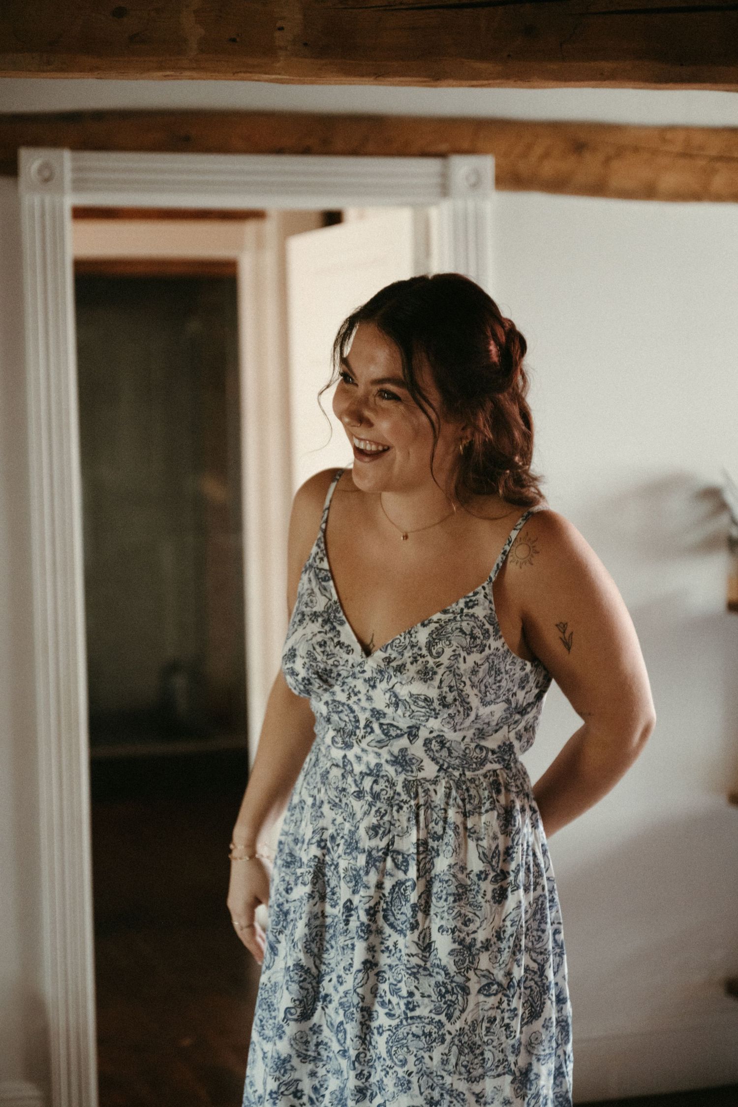 Young woman laughing in a blue floral sundress inside a rustic room with wooden beam ceiling.