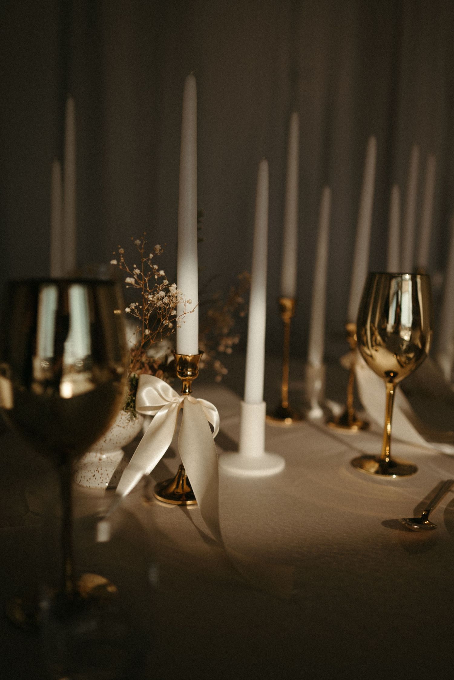 Elegant table setting with white taper candles in gold holders, gold wine glasses, and dried floral accents on a neutral linen.