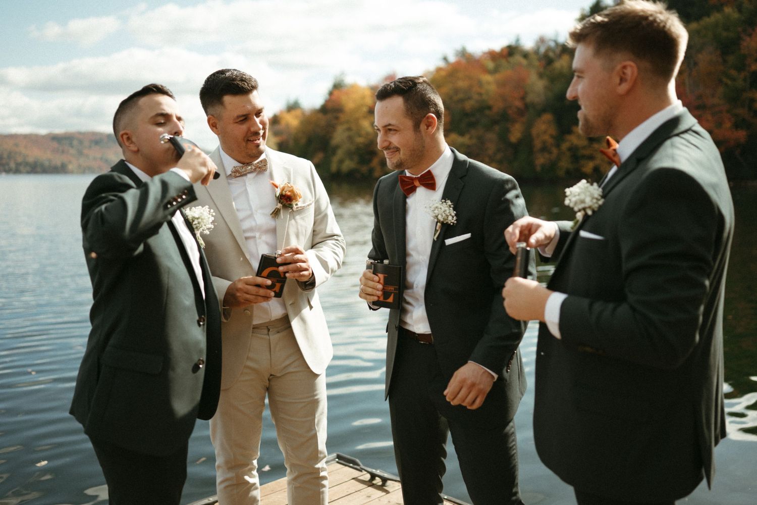 A group of four men in formal attire standing near a body of water on an autumn day.