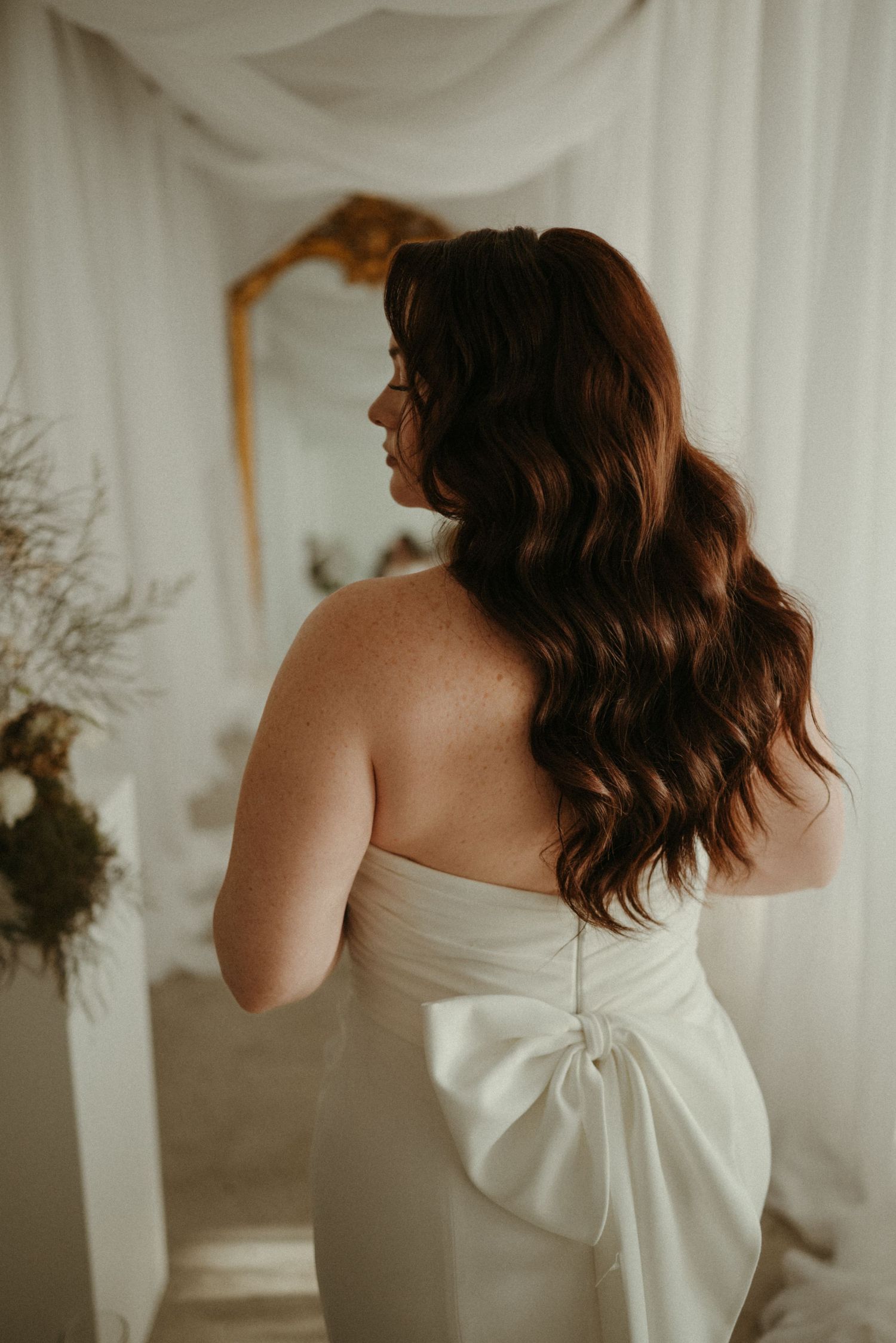 Bride in strapless white gown with large bow back, showcasing long wavy brunette hair in elegant bridal suite.