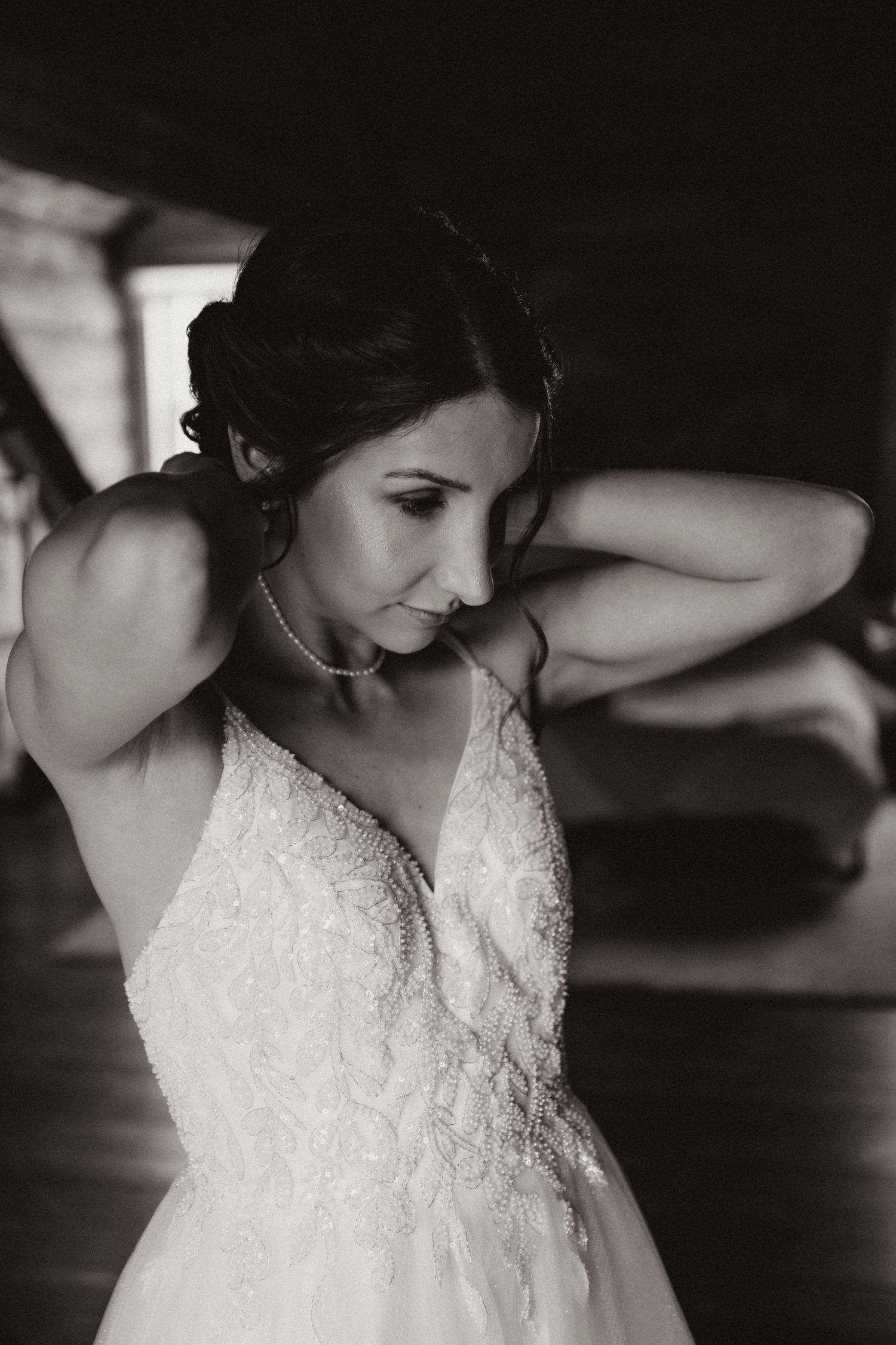 Black and white photo of a bride in lace wedding gown adjusting her pearl necklace before the ceremony.