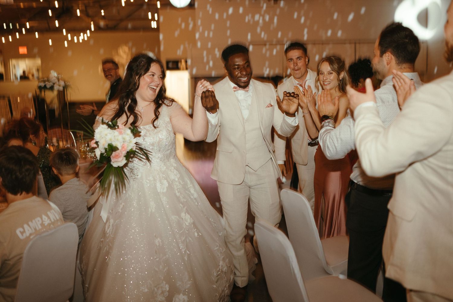Bride and groom walk joyfully through cheering guests at elegant indoor wedding reception celebration.