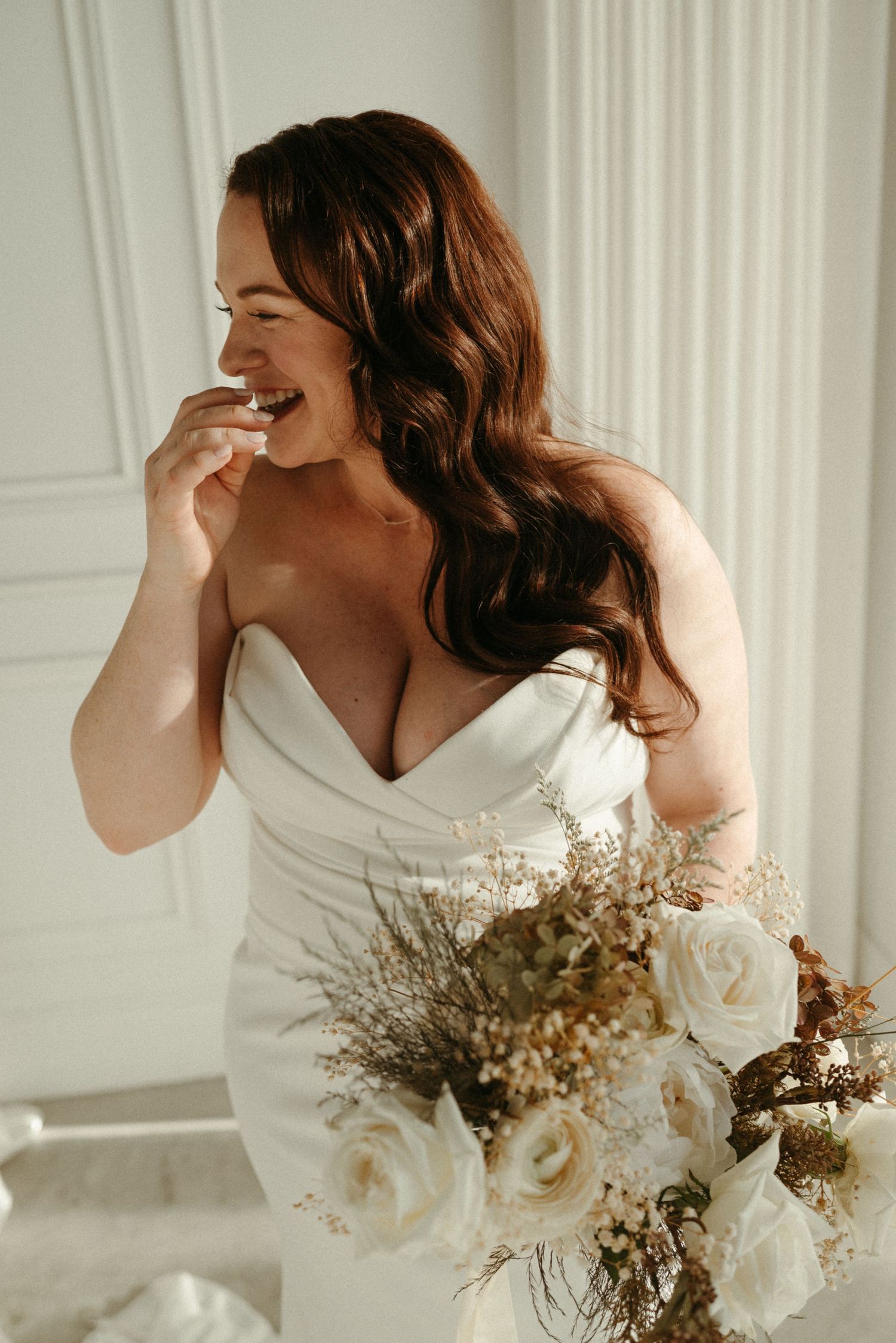 Laughing bride in strapless white gown holding bouquet of white roses and dried flowers in bright white room.