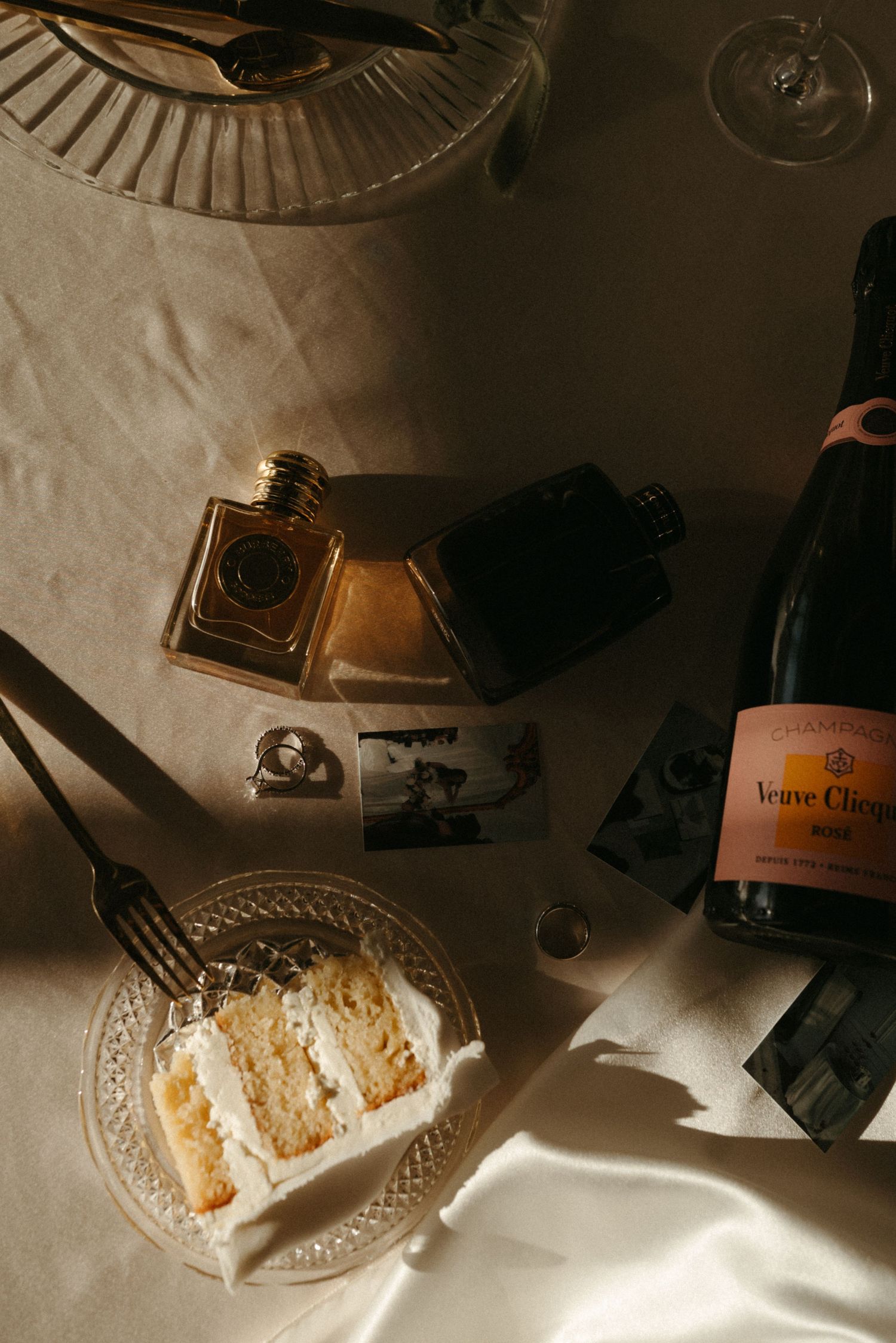 Flat lay of white wedding cake slice, Veuve Clicquot champagne, perfume bottles, and vintage photos on linen tablecloth.