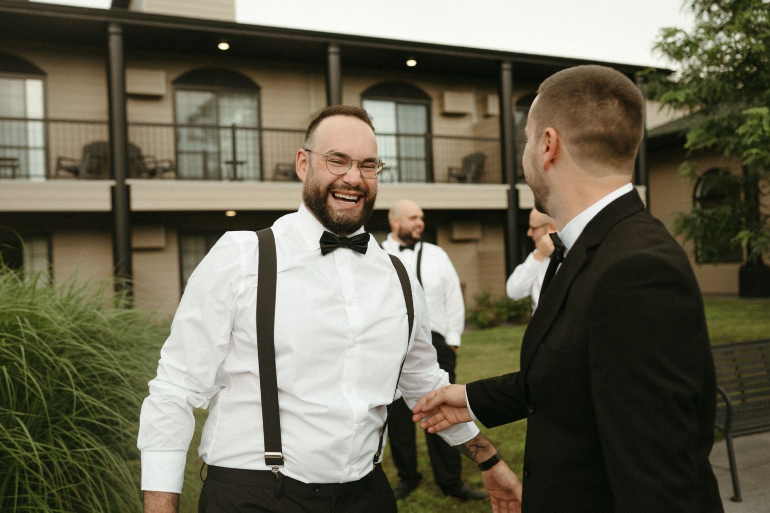 Groomsman in white shirt and suspenders shakes hands and laughs with groom in black tuxedo.