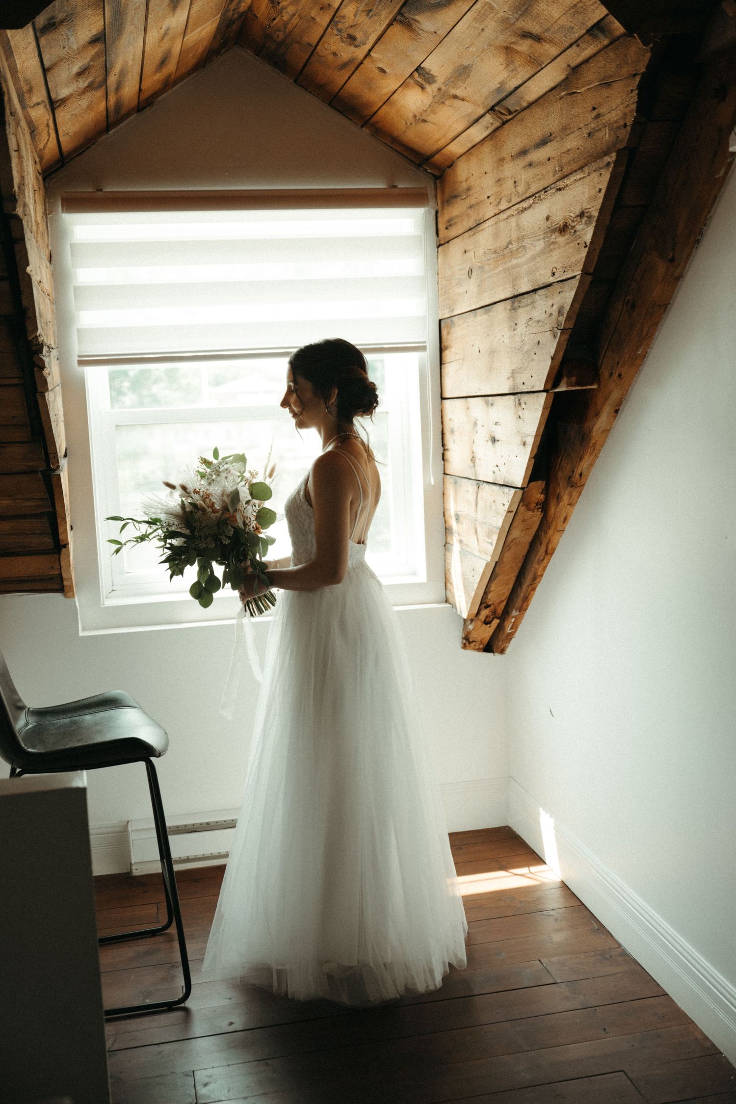 Bride in white tulle gown holding bouquet stands by attic window with rustic wood beam ceiling.