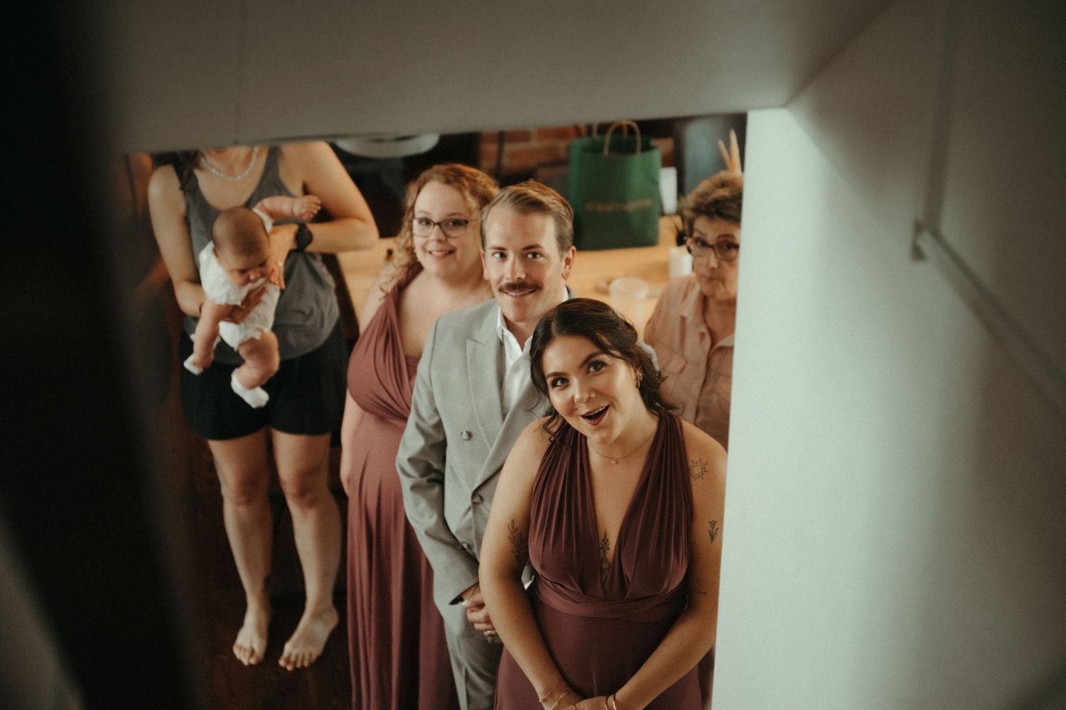 Wedding party in mauve bridesmaid dresses and gray suit gather in doorway, viewed from above at indoor venue.