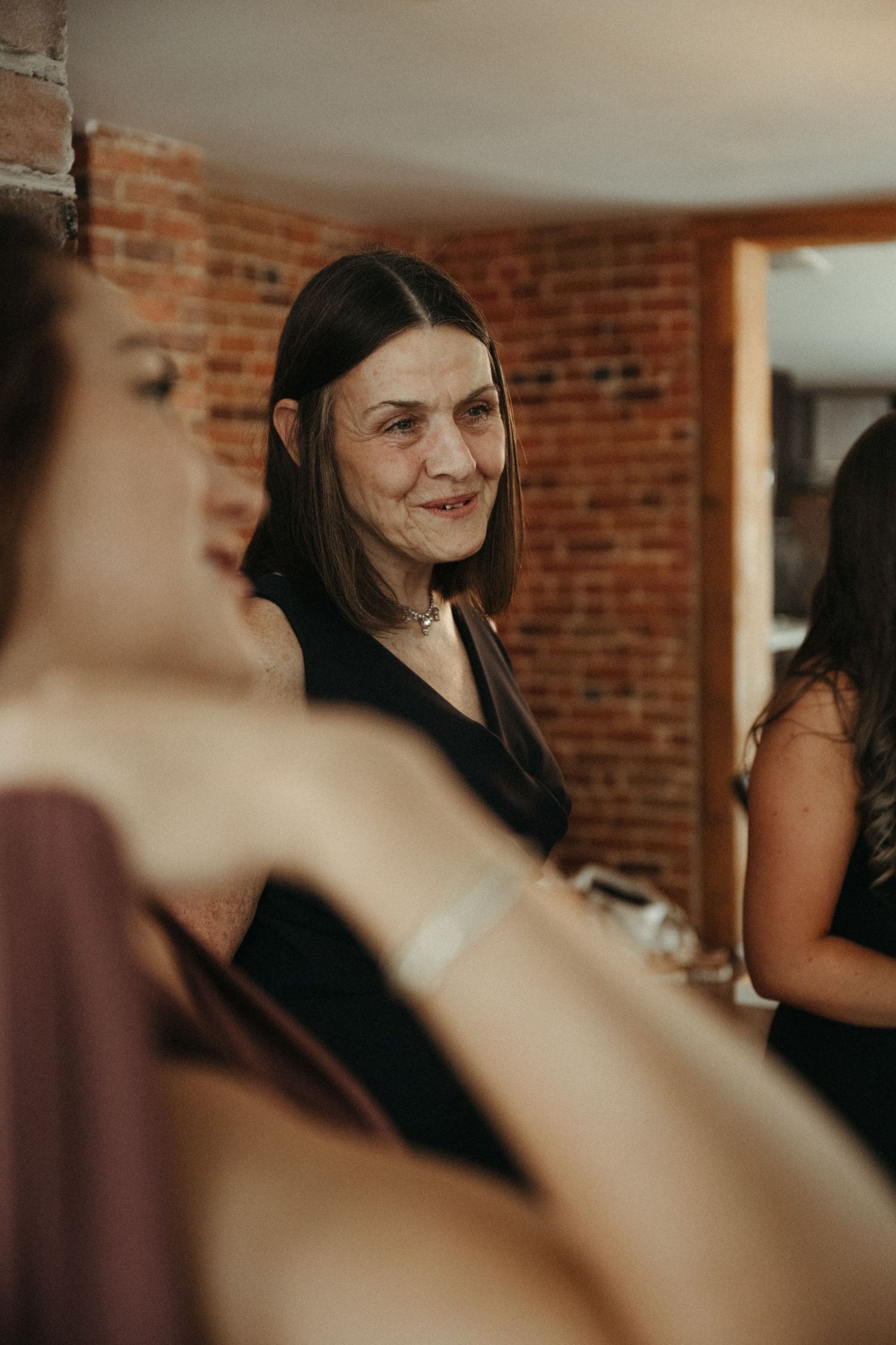Older woman smiling warmly at wedding guests in brick-walled venue with bridesmaids in foreground.