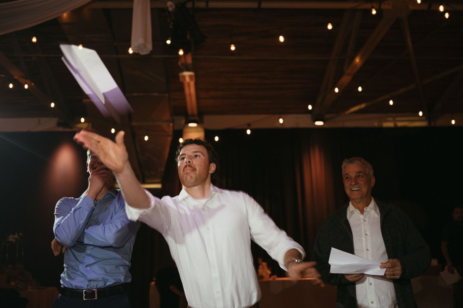 Man in white shirt tossing papers at an indoor event with string lights and guests in background.