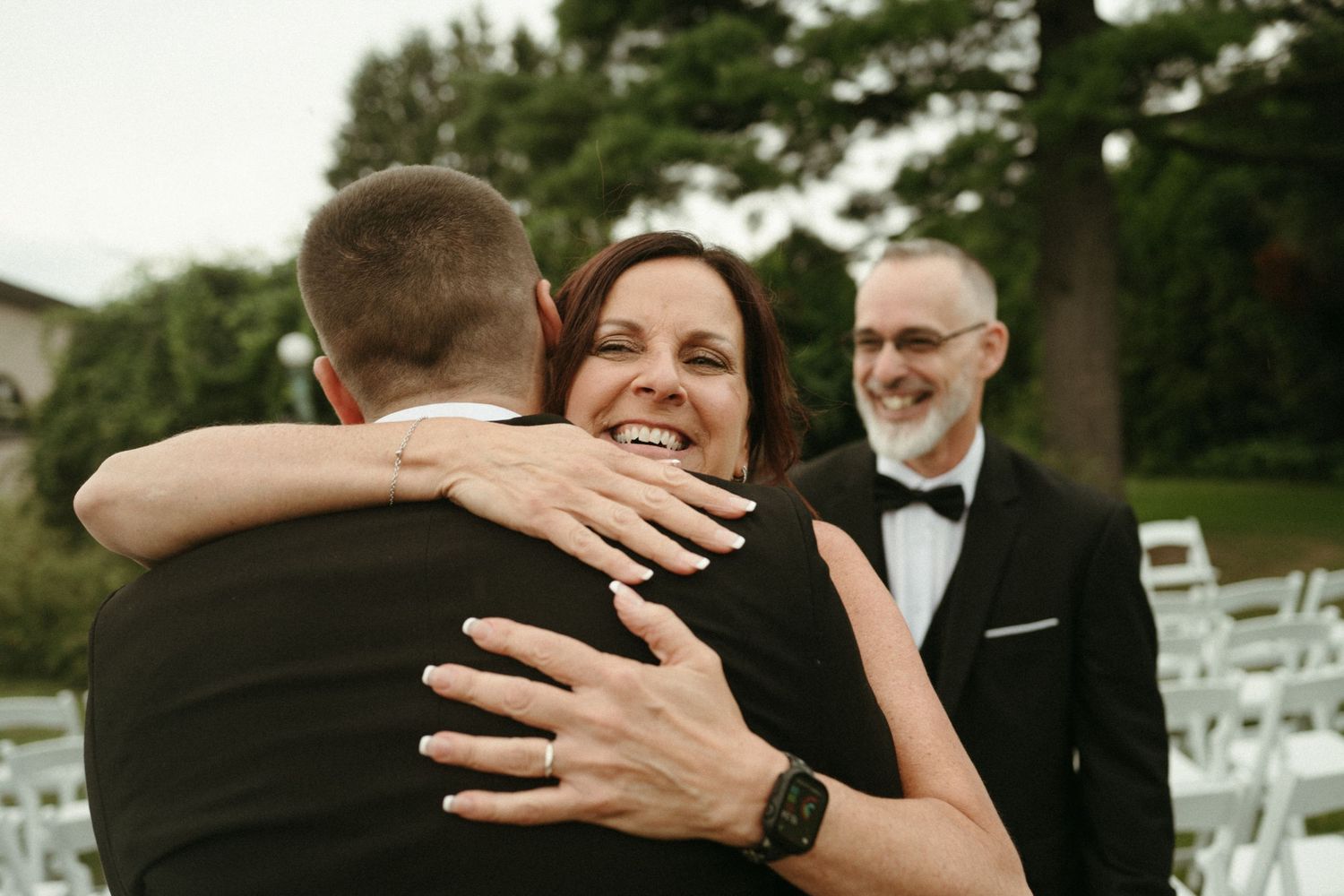Smiling bride hugs a guest in a tuxedo during an outdoor wedding ceremony.