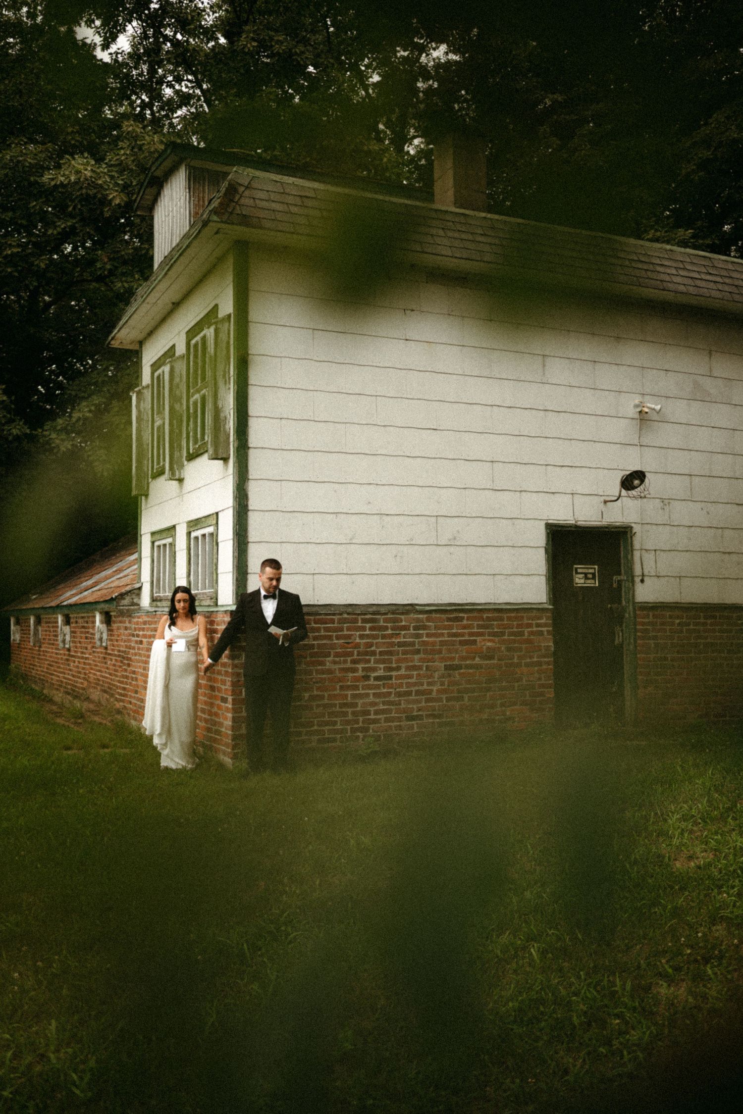 Wedding couple in elegant attire pose together against a white and brick building surrounded by lush greenery.