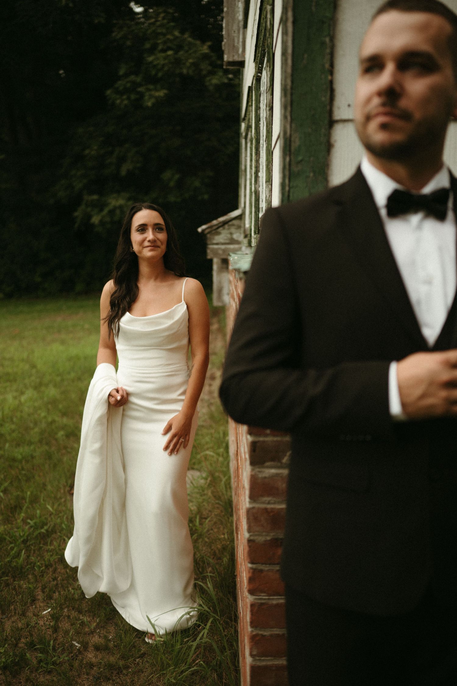 Bride in white gown stands by rustic brick building as groom in black tuxedo poses in foreground.