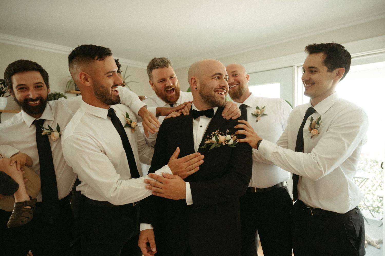 Groom in black bow tie laughing with groomsmen in white shirts and black ties before wedding.