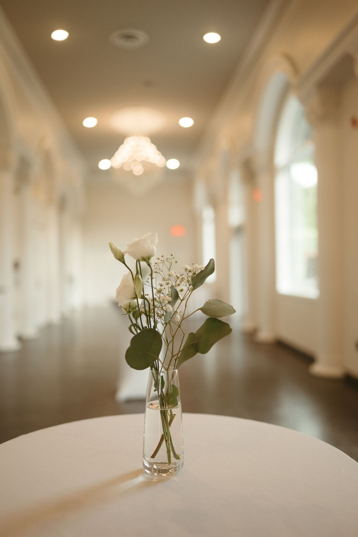 A glass vase with white roses and baby's breath on a white table in an elegant bright hallway with chandelier.
