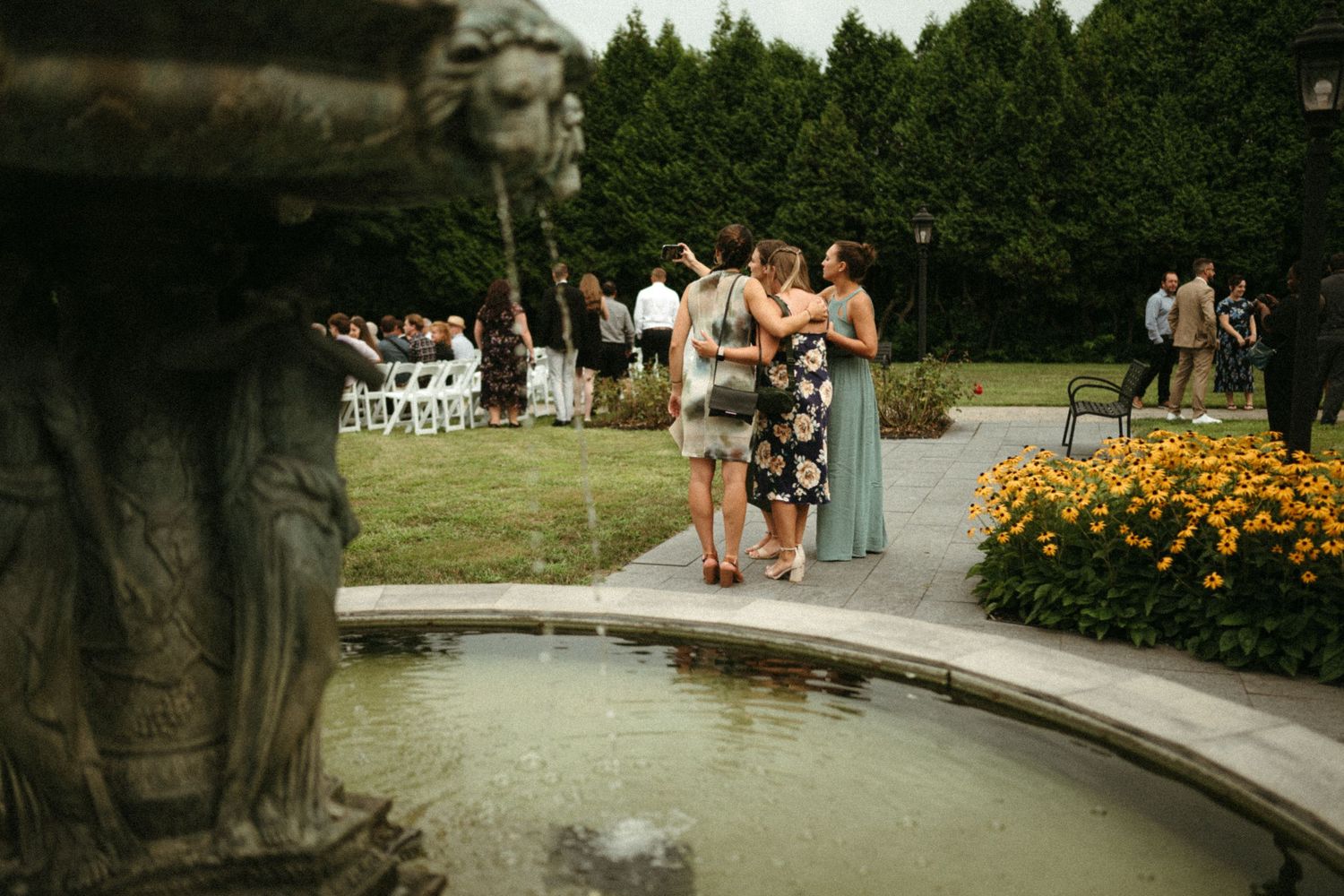 Wedding guests mingle near an ornate stone fountain during an outdoor garden ceremony.