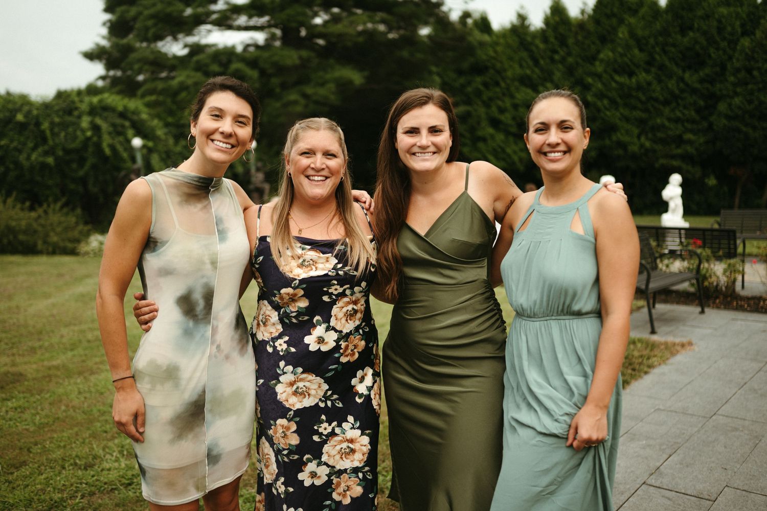 Four women in elegant dresses smile together at outdoor wedding reception garden venue.