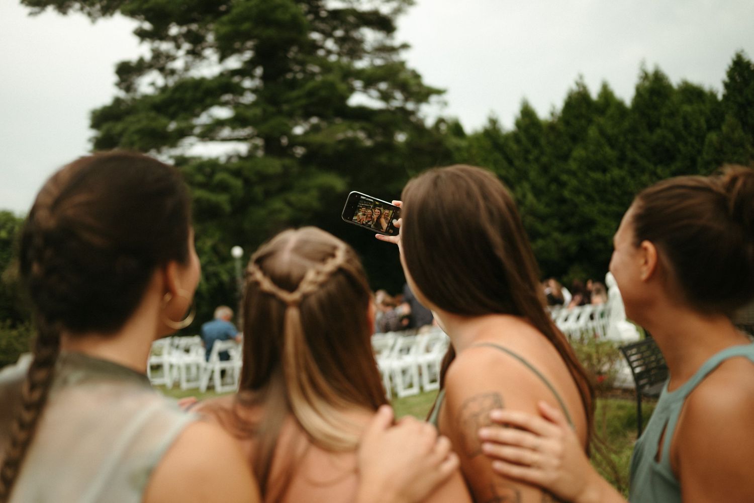 Wedding guests holding phones to capture outdoor ceremony with white chairs on lawn.