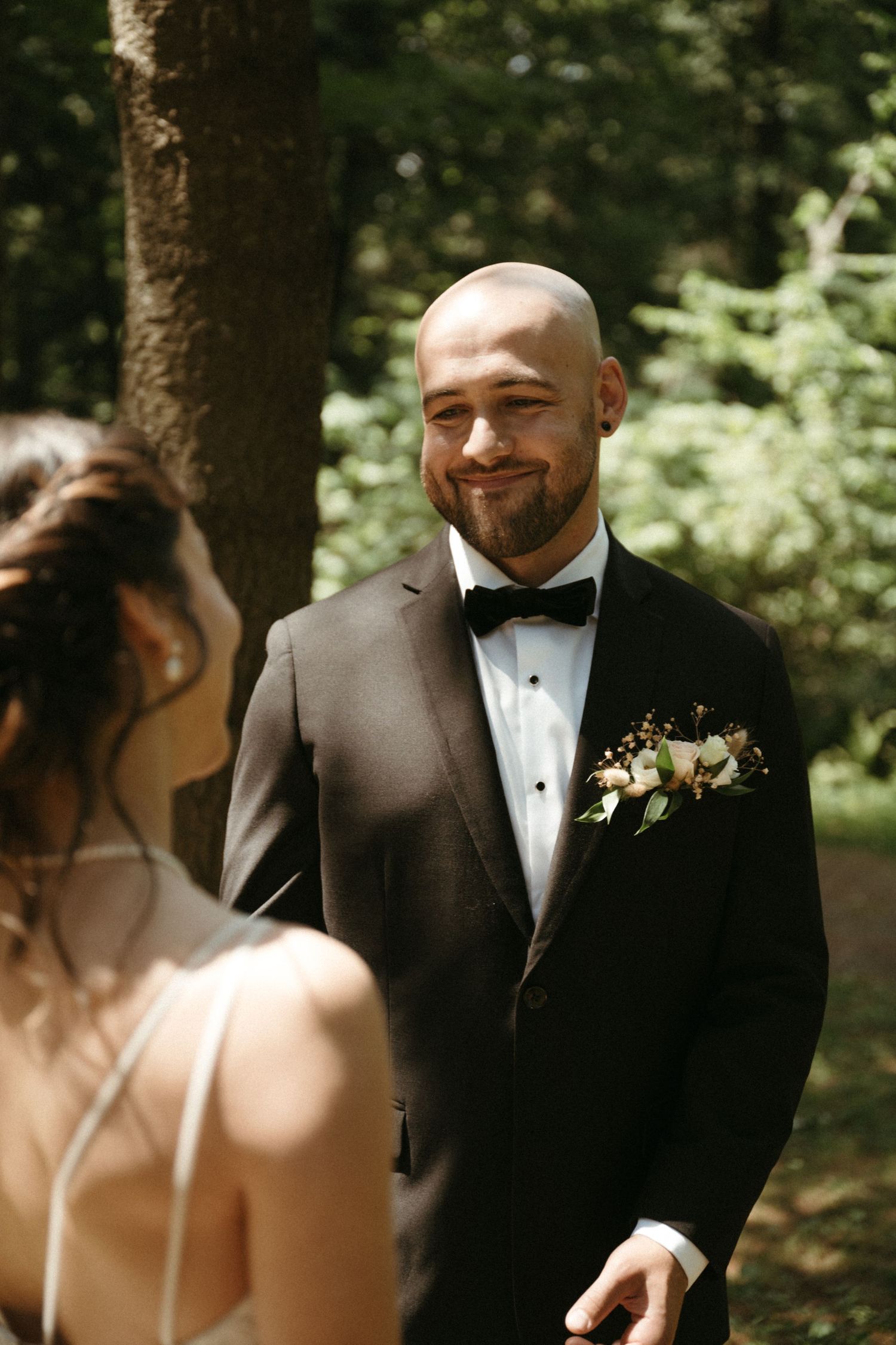Smiling bald groom in black tuxedo with floral boutonniere looking at bride during outdoor wedding ceremony.