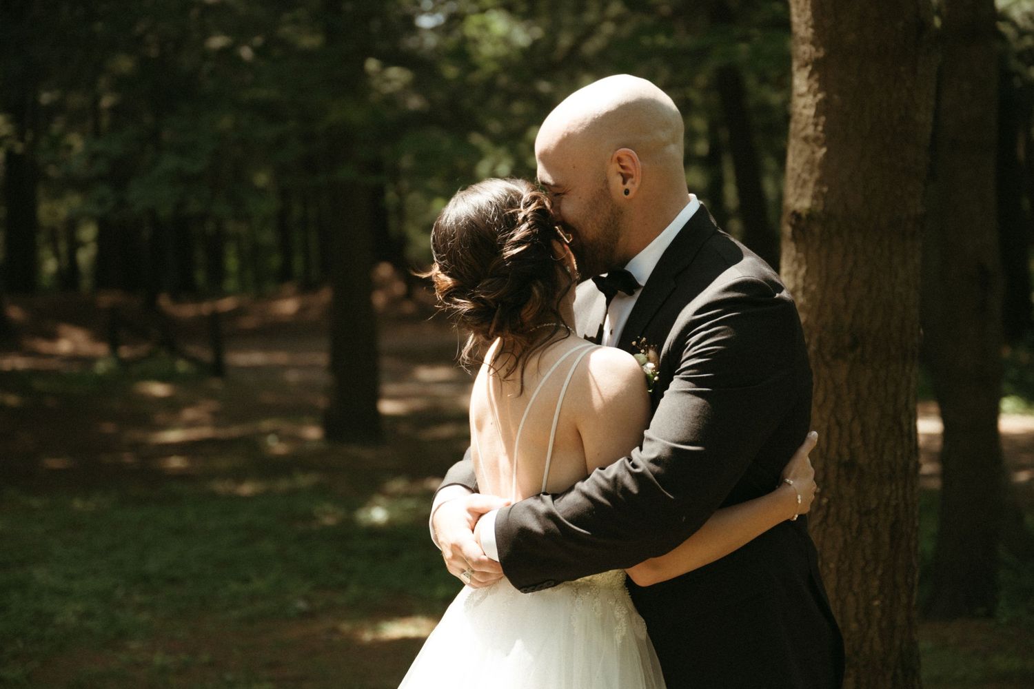 Bald groom in a black tuxedo sharing a tender moment with his bride in a forest.