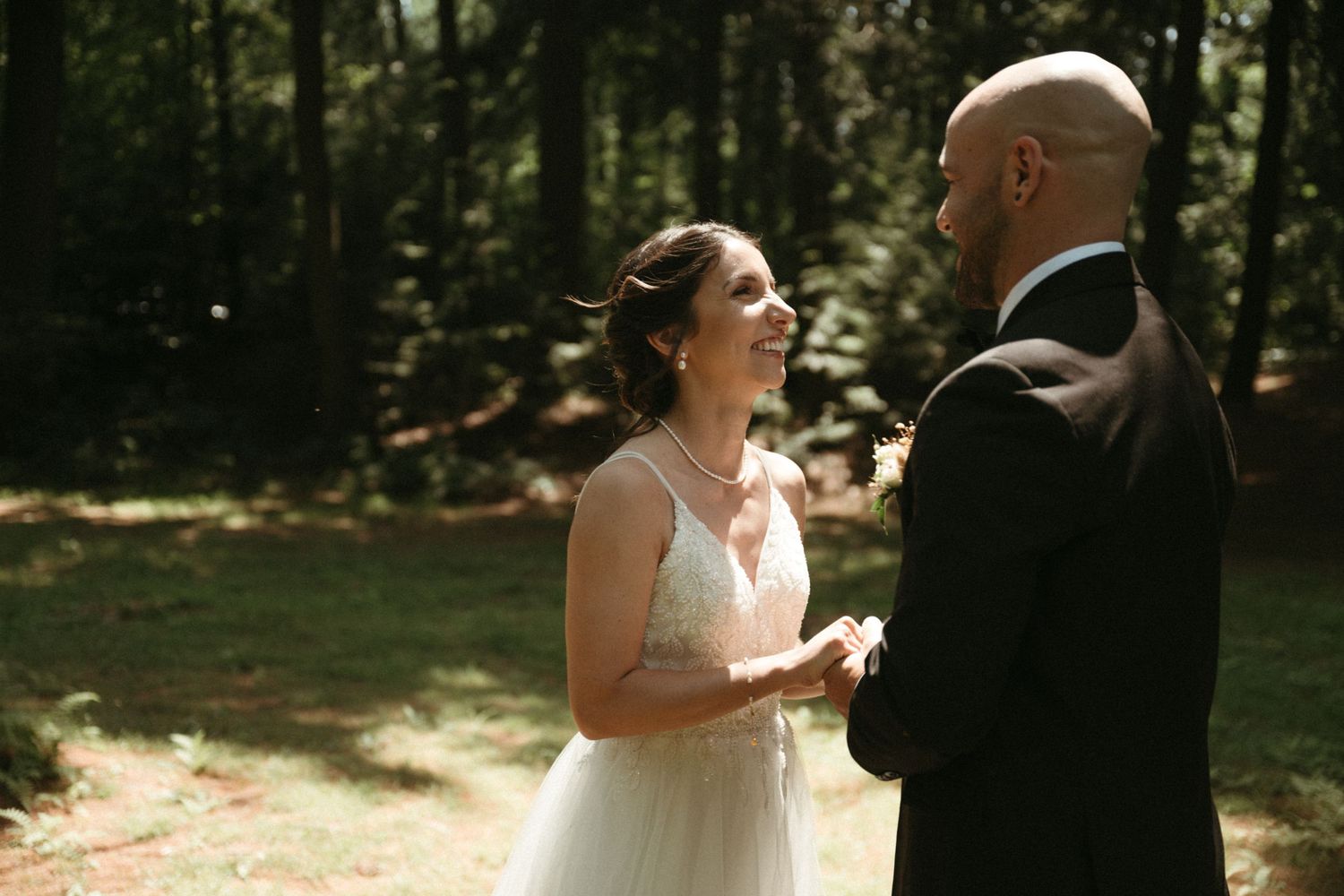 A smiling bride in a white gown shares a joyful moment with her groom in a forest setting.