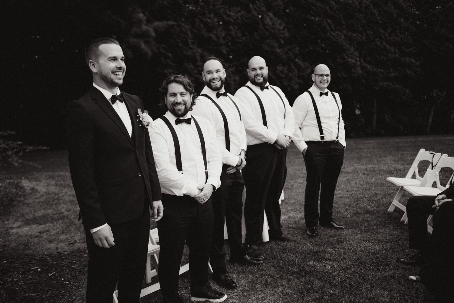 Groom and groomsmen in black tuxedos with suspenders posing outdoors in black and white photo.