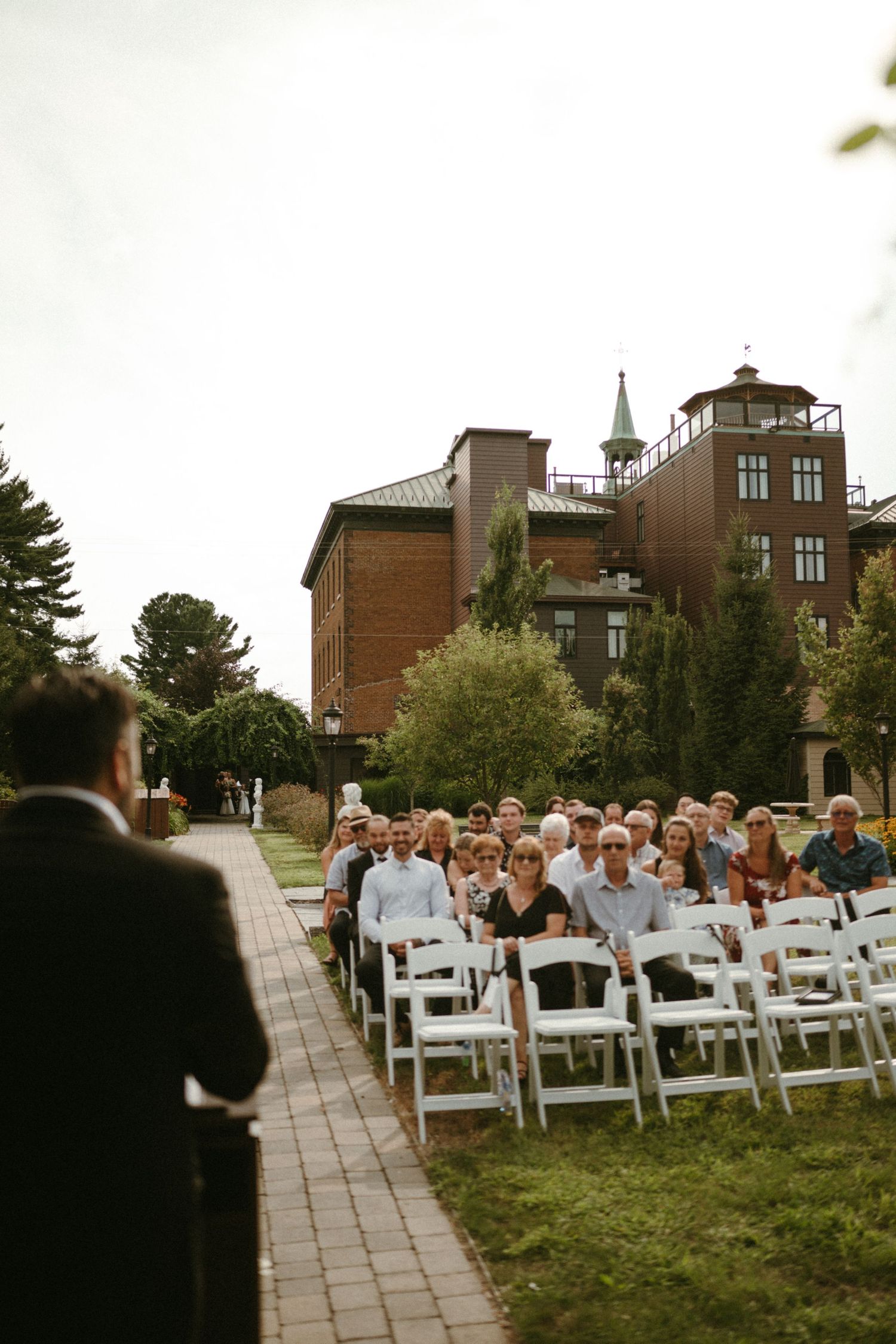 Wedding guests seated in white chairs outdoors facing the officiant with Hotel Montfort in background.