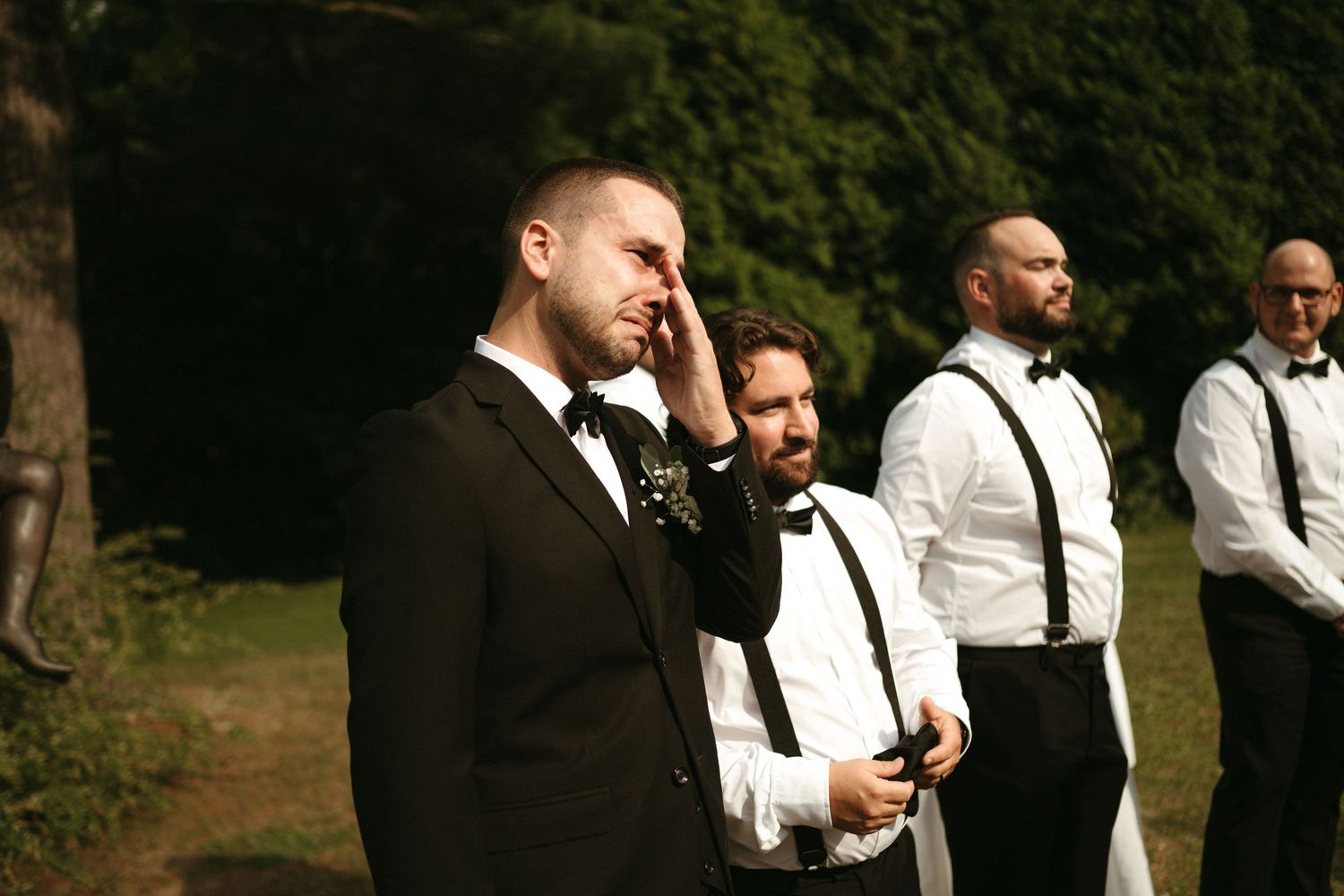 Emotional groom wipes tears while standing with groomsmen in white shirts and suspenders outdoors.