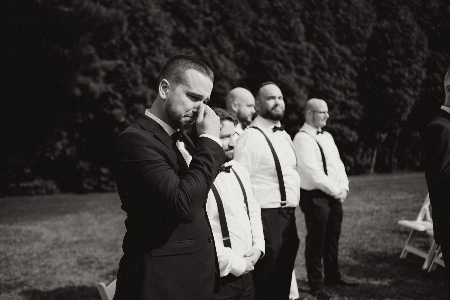 Emotional groom wiping tears with groomsmen in suspenders standing behind him during outdoor wedding ceremony.