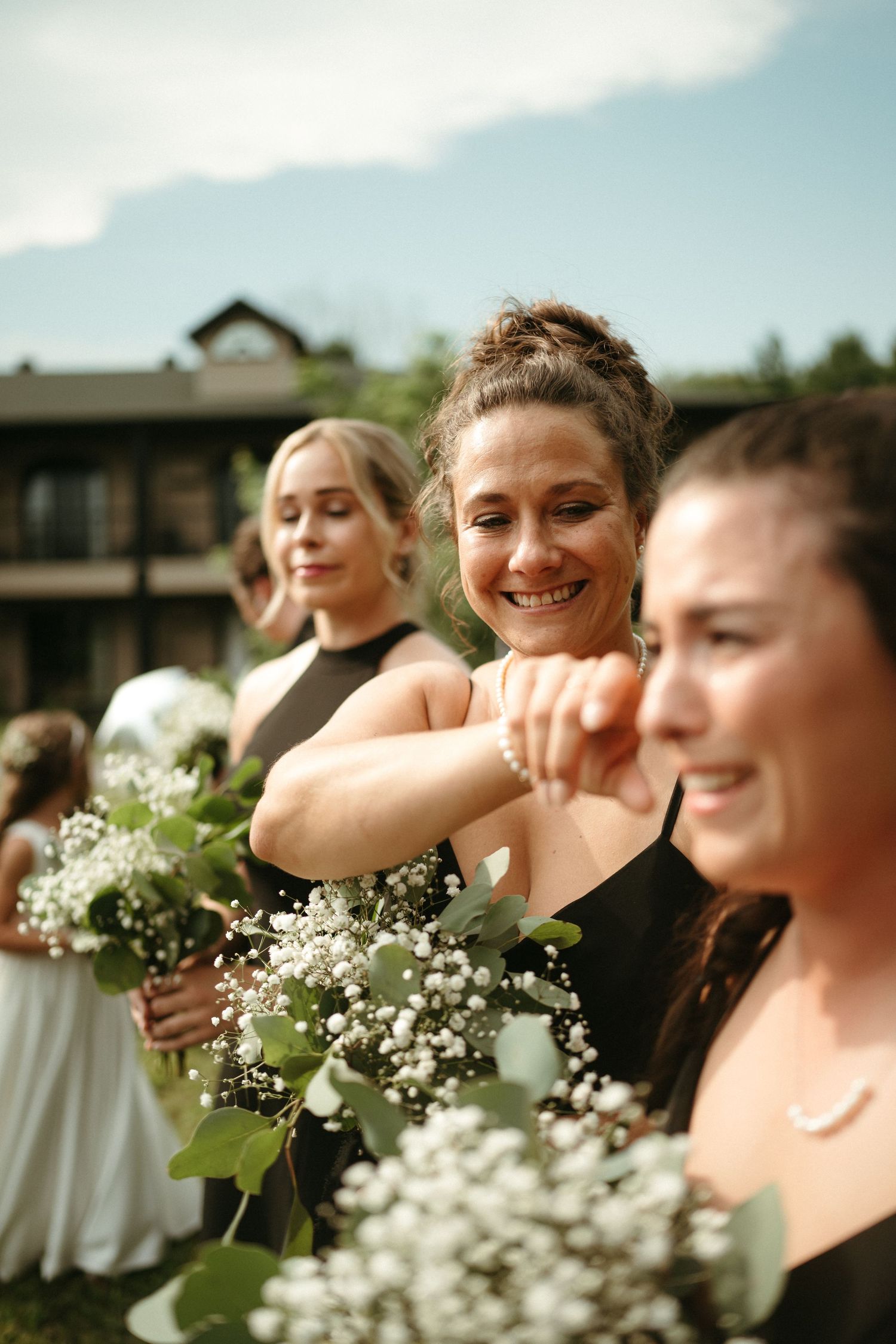 Bridesmaids in black dresses hold white floral bouquets with greenery outdoors at a wedding.