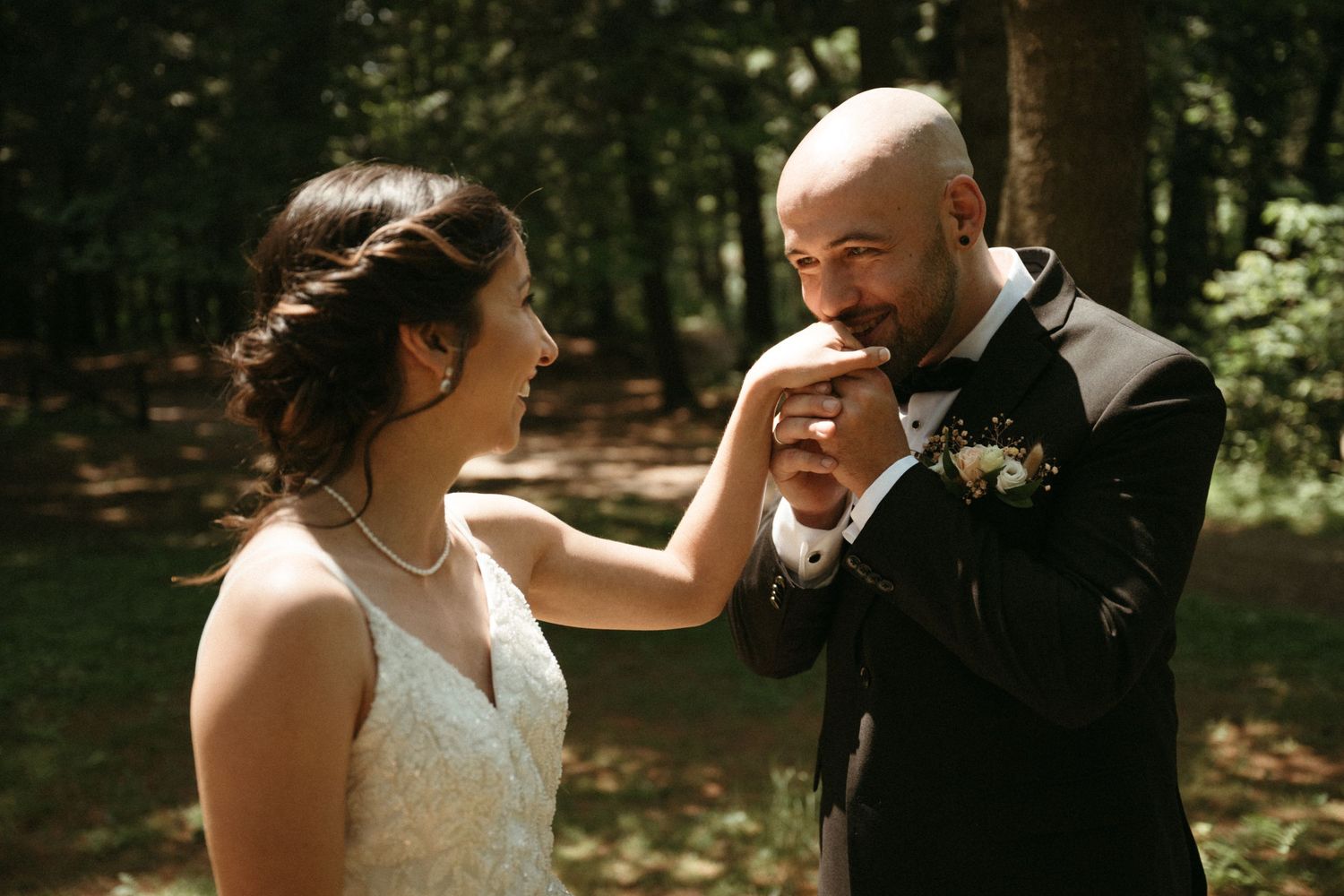 A bald groom in a suit tenderly kisses his bride's hand in a sunlit forest setting.